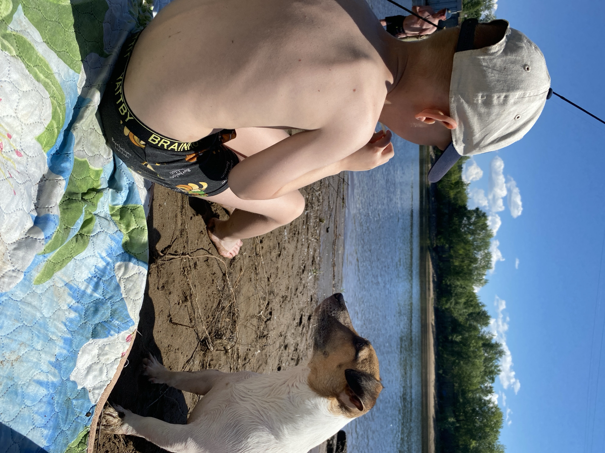 A boy in a beige baseball cap and black patterned swim trunks sits on a colorful blanket on the sandy bank of a river. He is facing away from the camera, looking towards the water. A small dog, primarily white with brown markings on its head and ears, sits to his right, also looking towards the river. In the background, a man in dark shorts stands in the shallow water. The river is wide, with a tree-lined bank visible across the water. The sky is blue with scattered white clouds. The scene appears to be a recreational area, possibly in the city of Chaikovsky, Russia, given the context of the request.