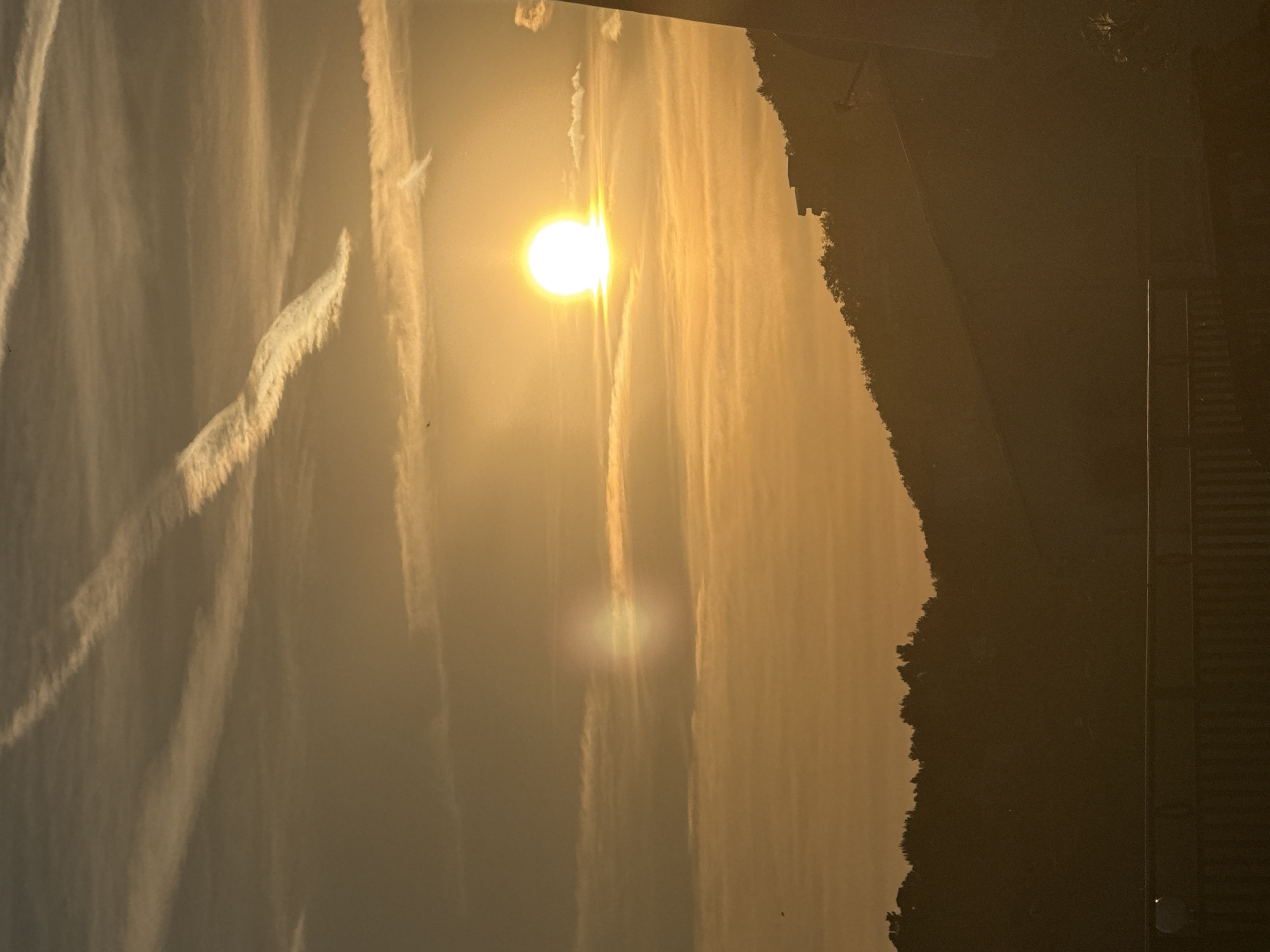 The image shows a sunset or sunrise in Šušanj, Montenegro. The sun is high in the sky, obscured by a light cloud cover, and surrounded by contrails. To the right of the sky is a dark tree-covered slope, and a portion of a building.