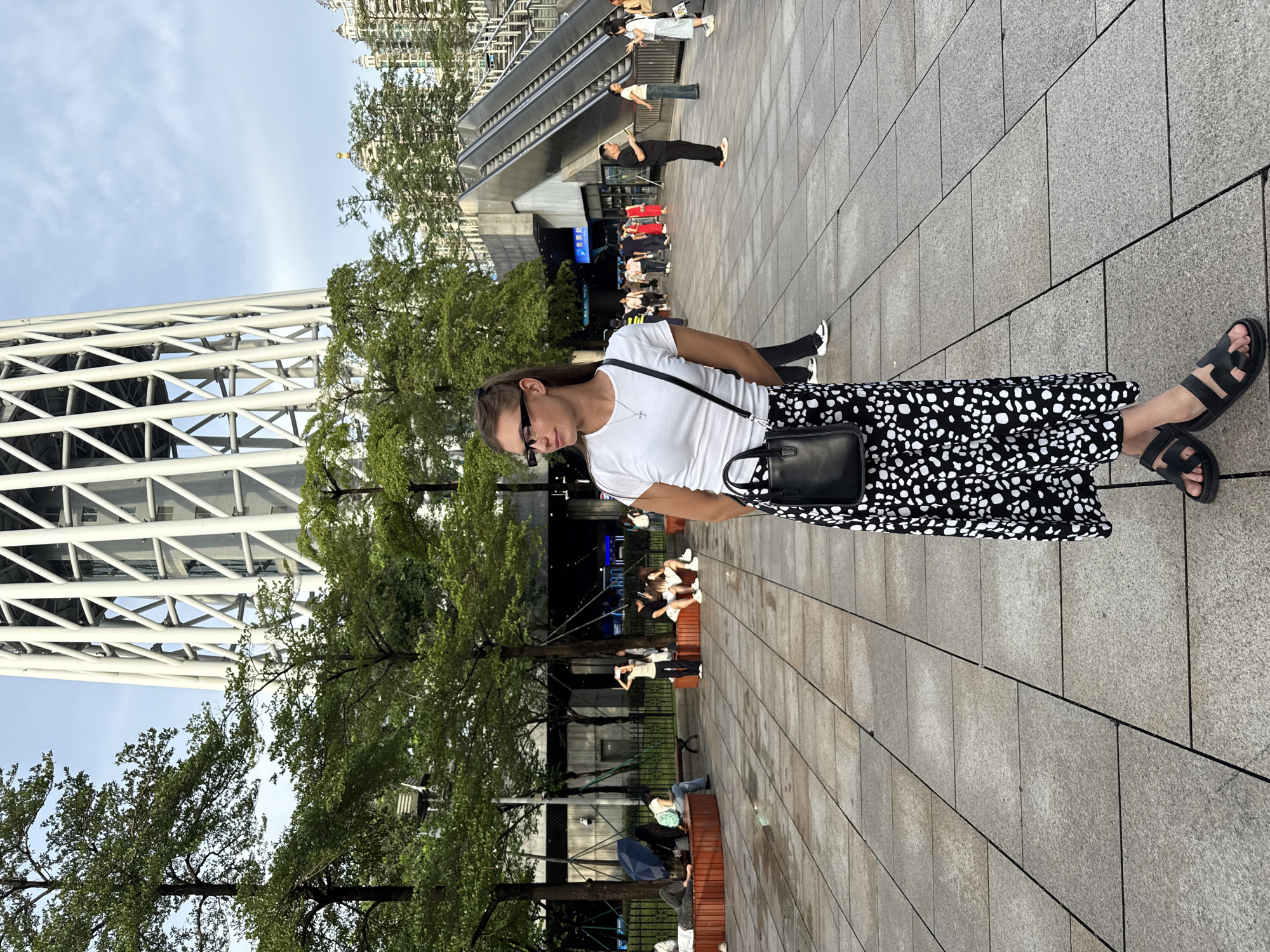 A woman stands in a paved plaza in front of a tall, white, lattice-like structure, likely a tower. She wears a white t-shirt, a black and white spotted maxi skirt, and black sandals, carrying a small black purse over her shoulder. She has on glasses and her hair is pulled back. Behind her, trees with green foliage are visible, as well as an escalator leading up to a building. Several other people are scattered throughout the plaza, some sitting on benches and others walking. The ground is paved with large, square tiles, some appearing wet. The sky is overcast. This scene is located in Haizhu District, China.