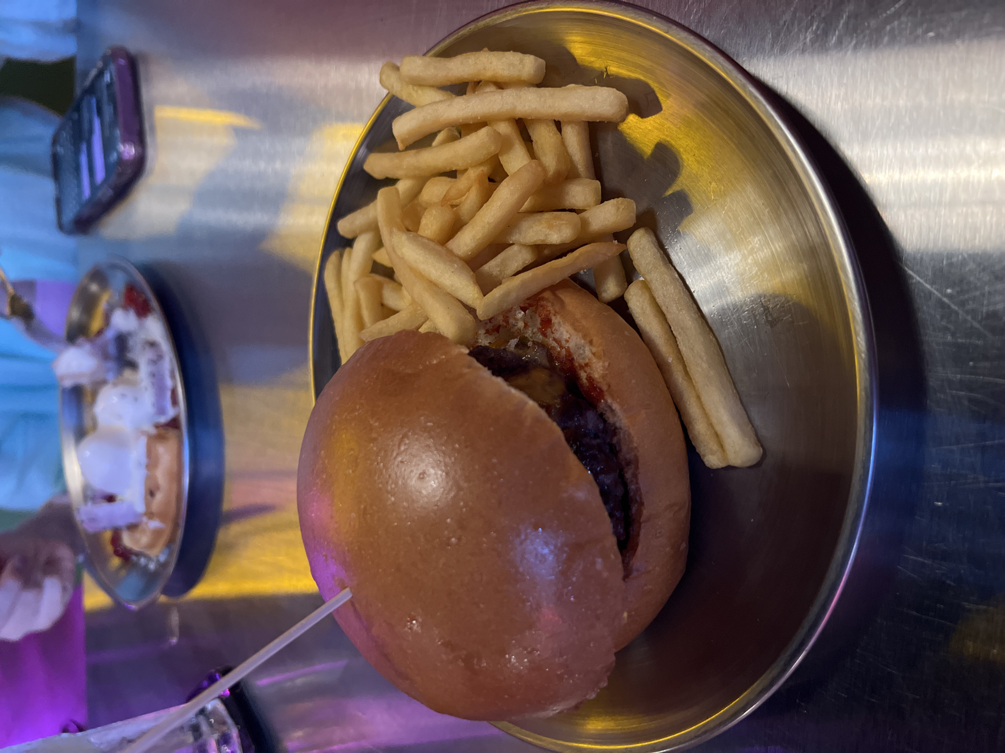 An overhead shot captures a casual meal served in a reflective, metallic bowl on a scuffed metal table. The main dish is a classic burger, held together by a white skewer, revealing a dark patty, melted cheese, and a reddish sauce, likely ketchup, nestled within a shiny, light brown bun. A generous portion of golden-brown french fries is piled next to the burger in the same bowl.
The metallic table surface and bowls reflect vibrant artificial lighting, casting prominent yellow, blue, and purple hues across the scene. These strong reflections suggest an indoor dining setting, possibly at night or in a venue with dynamic ambient lighting.
In the blurred background on the left, another small metallic bowl is visible, containing what appears to be a dessert with a creamy white substance and some reddish elements, possibly fruit or sauce. Further back, out-of-focus elements include a blurred purple object, possibly a smartphone, and hints of human presence like hands or arms, indicating an active dining environment. No text is visible in the image.