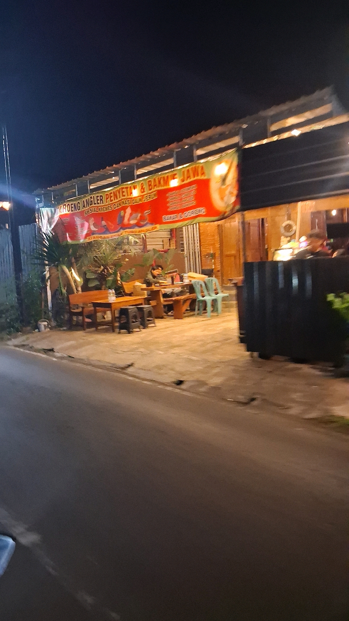The image captures an outdoor dining area of a restaurant at night, possibly located in Berbah, Indonesia, identified by a banner that reads "MROENG ANGLER PENYET & BAKME JAWA SPESIAL KREMES DAN NASI CULUN JERUK". The scene is illuminated by ambient lighting, casting long shadows.
In the foreground, a paved road or walkway is visible, with a dark, slightly reflective surface. To the right, a black, corrugated metal structure partially obscures the restaurant's facade.
The dining area features several wooden tables and benches. One table in the middle ground is occupied by a person sitting and looking down, possibly at a phone or food. Nearby, a few light blue plastic chairs are arranged around a table.
A prominent red banner with yellow and white lettering hangs across the front of the establishment, displaying the restaurant's name and menu items. Behind the tables, the restaurant building appears to have brick walls and wooden doors. Various plants and foliage are interspersed around the seating area, adding a touch of greenery to the night scene. The overall atmosphere is that of a casual, local eatery operating after dark.