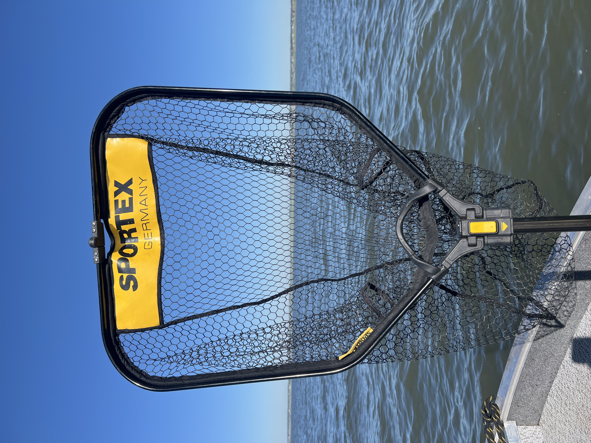 A fishing net, branded "SPORTEX GERMANY", is positioned in the foreground, obscuring the view of a body of water. The net's frame is black with a yellow label at the top. The net itself is black mesh. The water appears choppy under a clear blue sky. The lower edge of a boat, made of aluminum, is visible at the bottom of the frame. The location is specified as the city of Novopichugovo, Russia, however, this information is not visually verifiable from the image alone. There are no people or animals present in the frame.