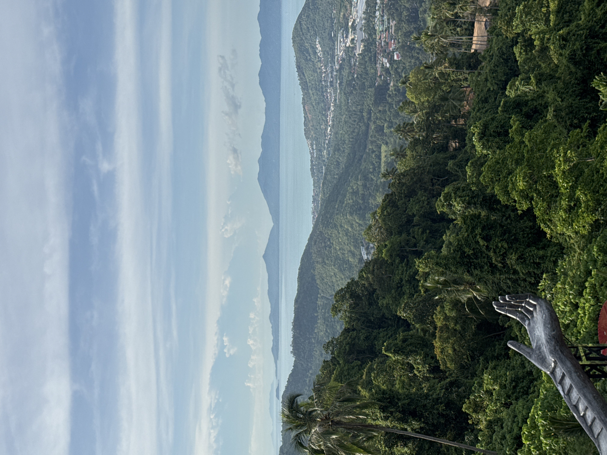 An elevated, wide shot captures a verdant landscape of rolling, densely forested hills meeting a calm, blue sea under a sky with scattered clouds. In the foreground, a large, metallic sculpture of an open hand with outstretched fingers is visible on the left, appearing to emerge from a dark, weathered structure. Beyond the foreground, the lush green hills are dotted with palm trees and other tropical foliage, descending towards a coastal town with buildings nestled amongst the trees and along the shoreline. In the distance, several mountain ranges fade into the hazy horizon, with the sea stretching out before them. The lighting suggests it is daytime, with soft sunlight illuminating the scene. The location is described as Unknown, Unknown.