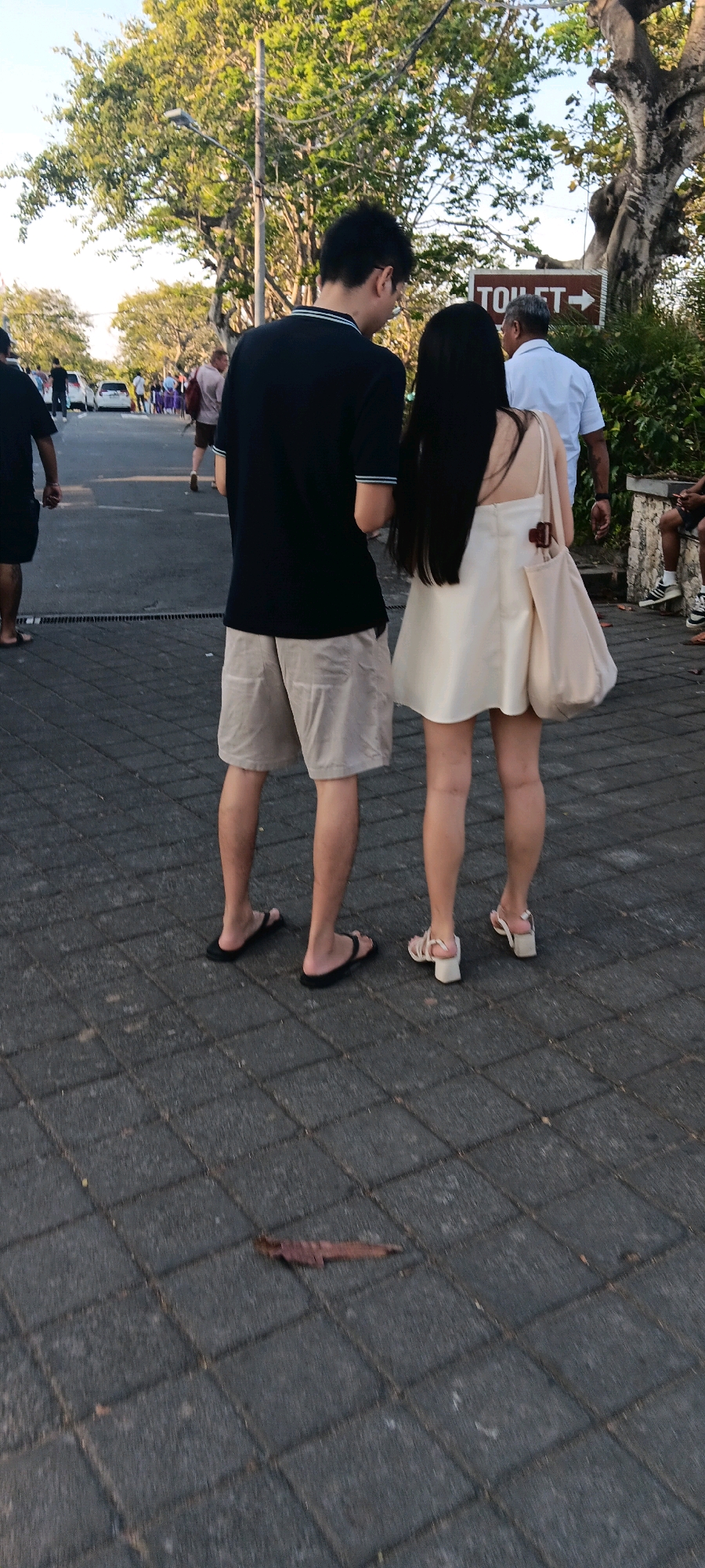 In Kuta Selatan, Indonesia, a couple stands on a paved walkway. The man, wearing a black polo shirt and beige shorts, is facing away from the camera, while the woman, in a cream-colored dress, stands beside him, also facing away. A large, light-colored tote bag hangs from her shoulder. In the background, a sign points to a toilet. People and vehicles are visible on a road in the distance. Trees with lush green foliage line the area, and the ground is a mosaic of gray square pavers.
