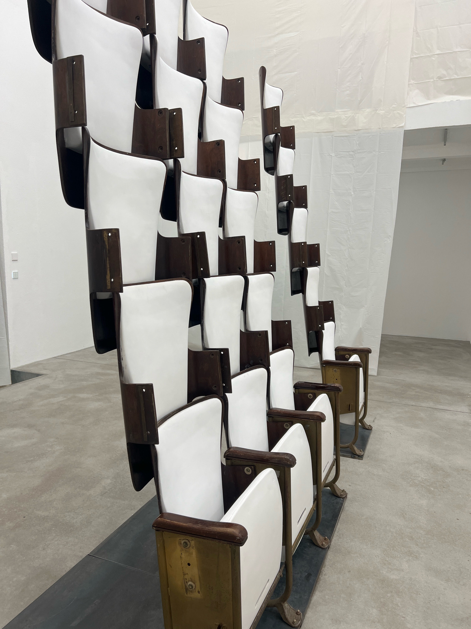The image depicts an art installation featuring a tiered arrangement of old theater seats. The seats have white plastic or wooden backs and seats, with dark wood trim and metal frames, some of which appear aged and patinated. The installation is set against a plain white background, likely a gallery wall, with a white fabric or tarp covering the upper portion of the wall on the right side. The floor is a concrete surface. The arrangement of the seats is staggered and stacked, creating an abstract, sculptural form. The lighting is even, suggesting an indoor exhibition space. The specific location in Berlin, Germany, is not identifiable from the visual information alone. There are no people present in the image.