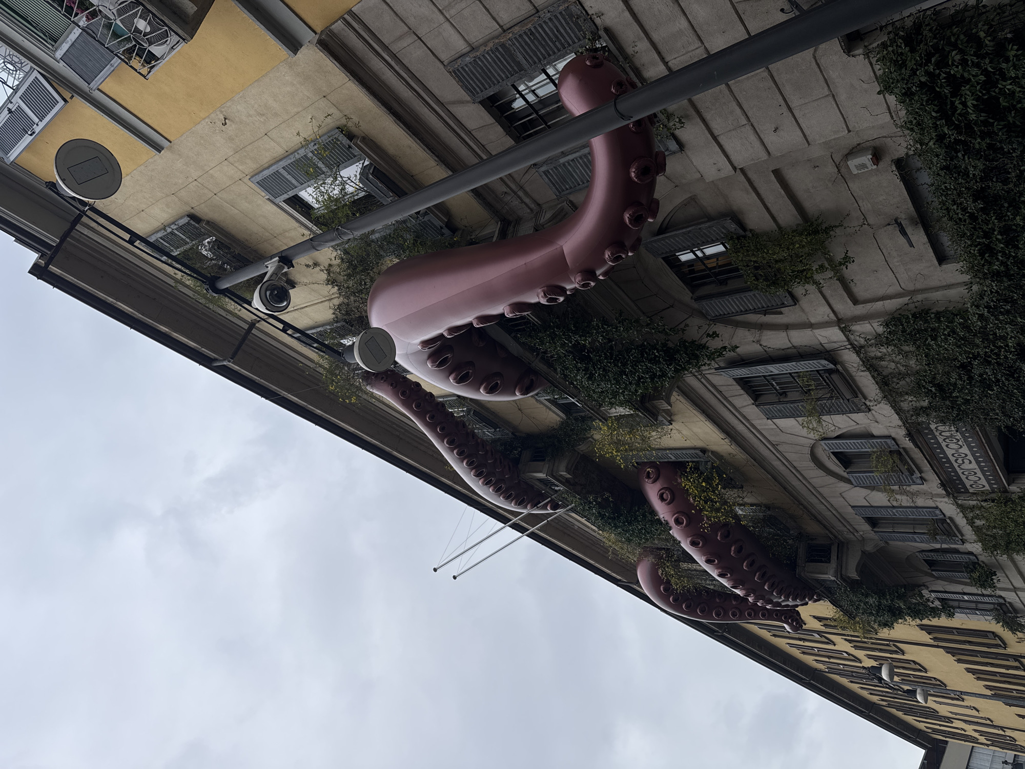 The image shows a building in Milan, Italy, decorated with large, inflatable octopus tentacles protruding from the windows. The building has multiple windows with shutters, some overgrown with greenery. A security camera is visible. Streetlights and poles are attached to the building's facade. The sky is overcast.