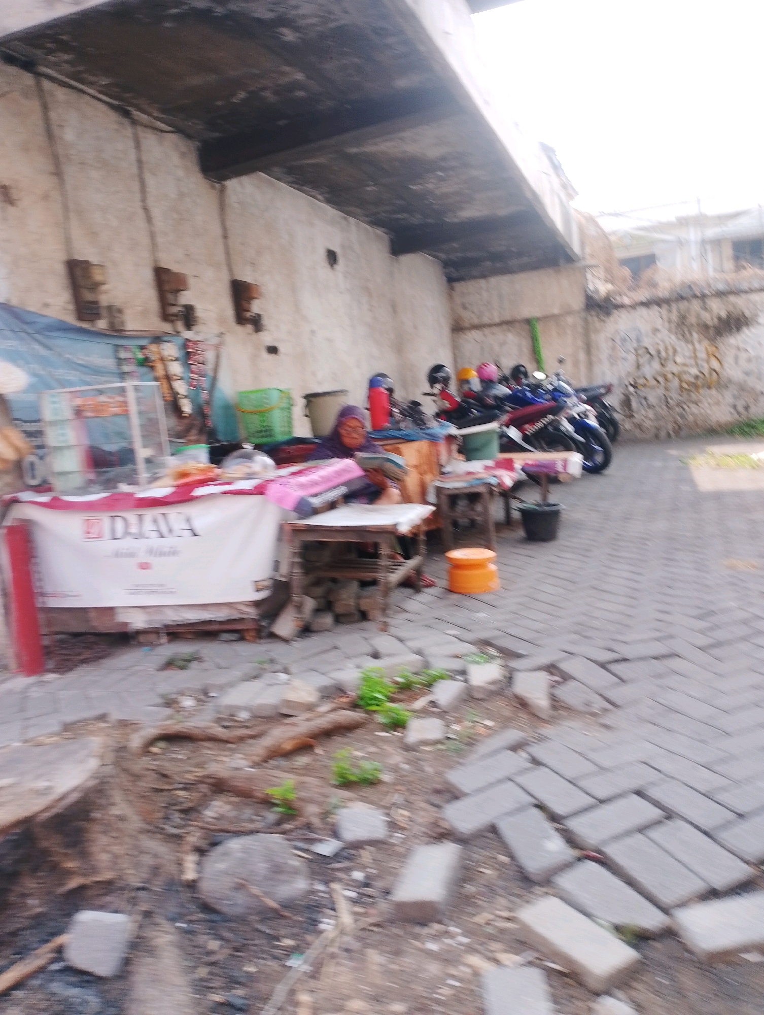 The image depicts an outdoor scene in Probolinggo, Indonesia, likely in a market or roadside stall area. A woman wearing a purple headscarf is seated behind a stall, which displays a banner with "DJAVA" written on it. The stall has a glass display case and various items, including what appear to be packaged goods and a pink tray. To the right of the stall, a wooden table with a small orange stool and a black bucket is visible. In the background, several motorcycles are parked, suggesting transportation is a common element in this location. The ground is paved with interlocking bricks. The overall environment appears somewhat rustic, with exposed concrete structures and some debris on the ground. The lighting suggests it is daytime, with a bright sky.