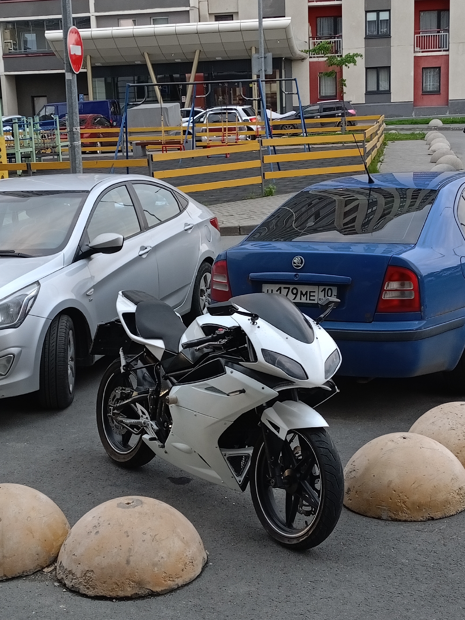 A white motorcycle is parked on asphalt in front of a blue Skoda Octavia, which has the license plate "179ME10". To the left of the motorcycle is a silver Hyundai Solaris. In the background, a playground structure with yellow and blue elements is visible, along with a red and white building. The scene appears to be in a residential area, possibly a courtyard or parking lot. The overall impression is a street-level view of parked vehicles and urban infrastructure.