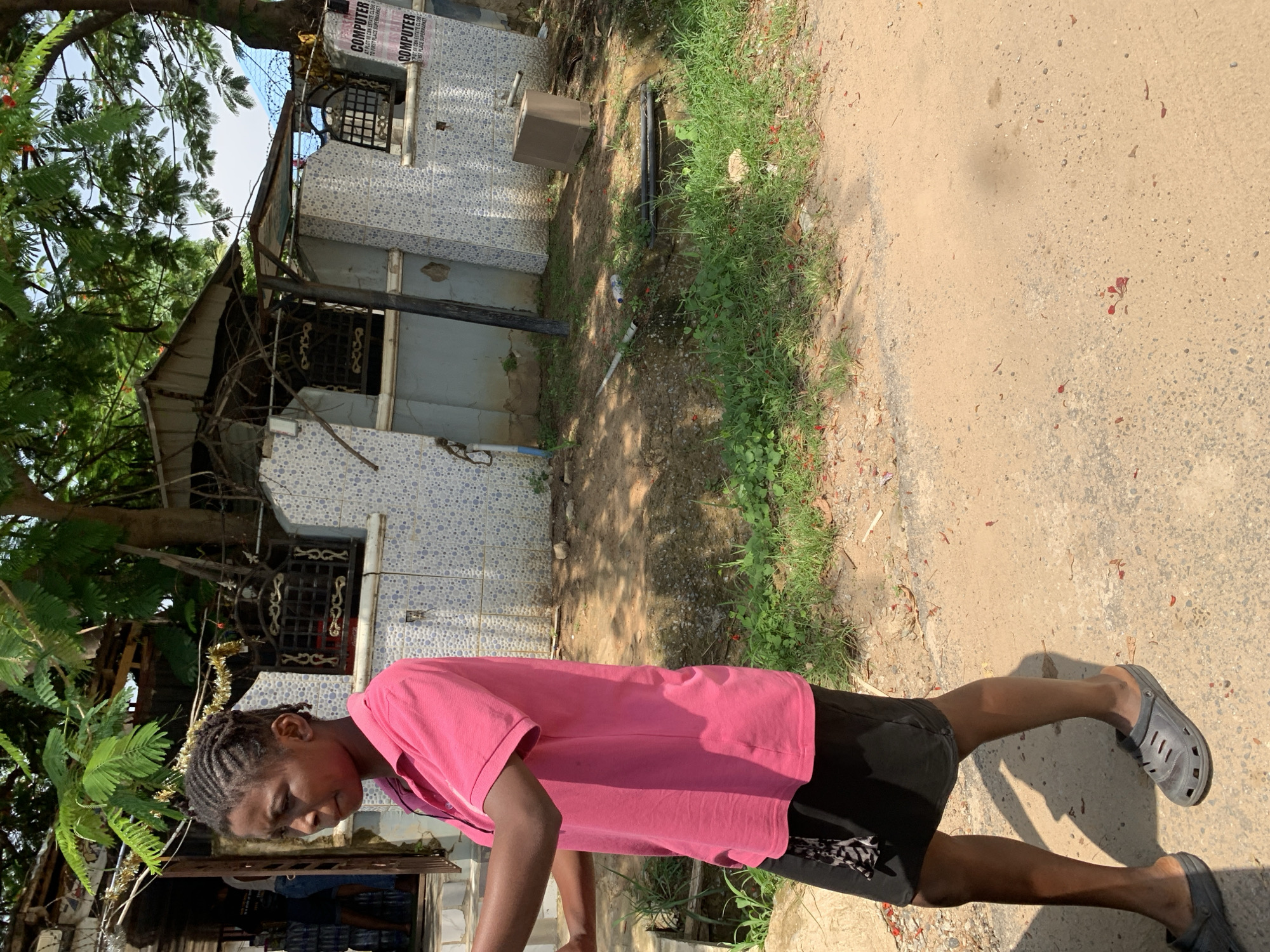 A young girl with braided hair, wearing a pink polo shirt and a black skirt, walks on a dirt road in Abuja, Nigeria. Behind her, a building with light blue and white patterned tiles has a corrugated metal roof and barred windows. Trees with green leaves and some red flowers overhang the scene. To the right of the building, two posters with text are visible. The ground is a mixture of dirt, sand, and sparse green grass.