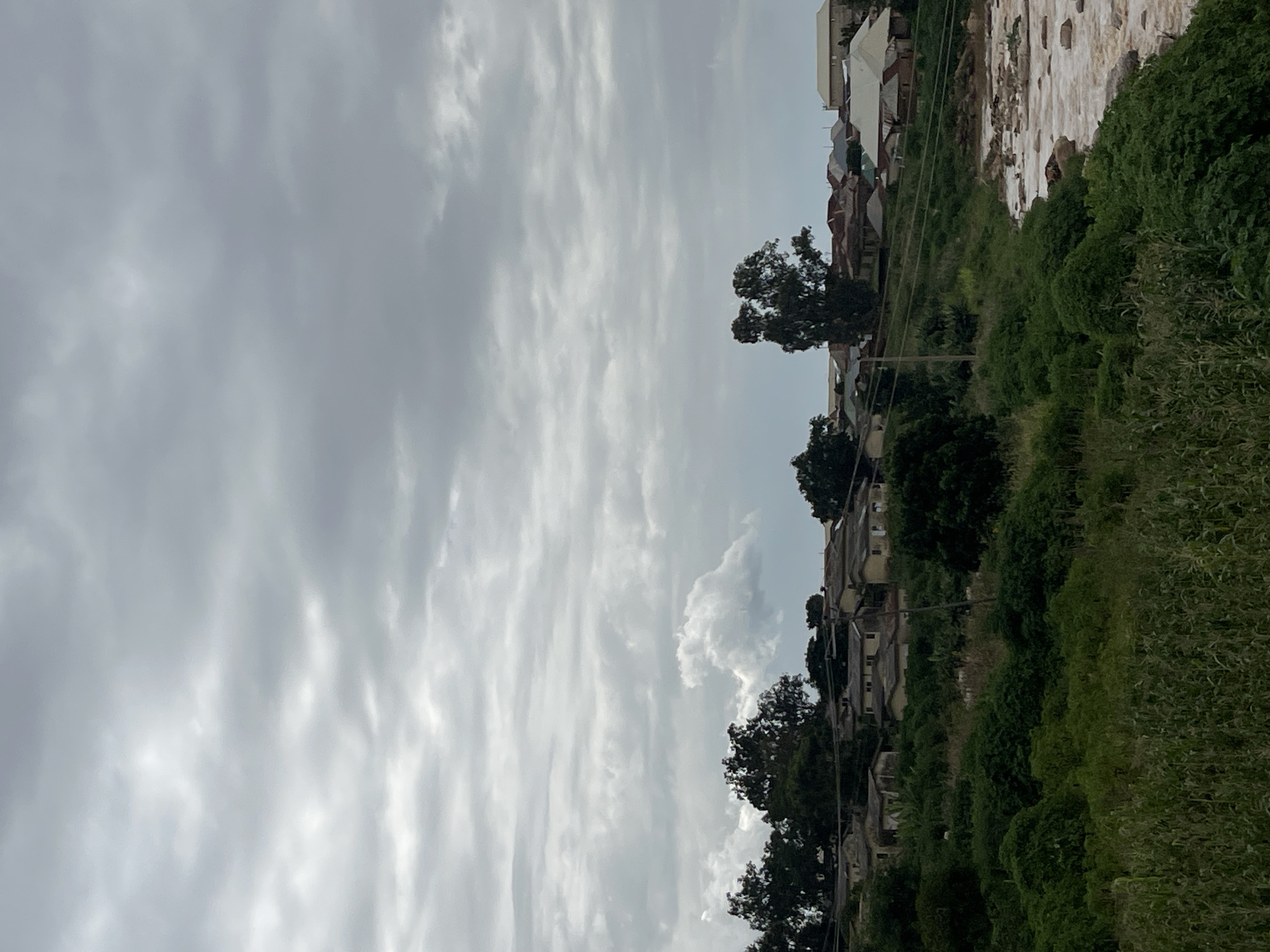 The image shows a rural hillside settlement under a cloudy sky. The settlement consists of numerous single-story houses with visible rooftops, situated on a sloping terrain covered with dense green vegetation. Trees are interspersed among the houses and on the hillside. A body of water with visible rocks is on the right side of the image, bordering the vegetated area. The overall scene suggests a somewhat undeveloped area with a visible natural water feature. The location is identified as Rafin Par, Nigeria.