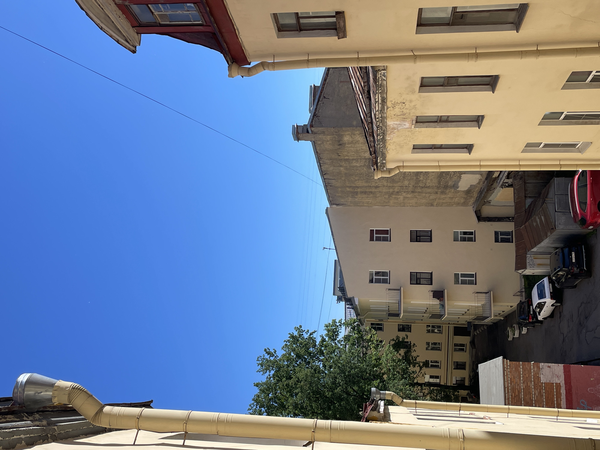 The image depicts a view of a courtyard in Saint Petersburg, Russia, on a clear, sunny day. The viewpoint is elevated, looking down into the courtyard area and at the surrounding buildings.
In the foreground, on the left, is a section of a light yellow building with a prominent downspout running vertically along its side. Adjacent to this, a lower structure with a red and white brick facade is visible.
The courtyard itself is paved and narrow, with several cars parked along its length. A white sedan, a black SUV, and another black car are clearly visible, along with a red hatchback parked at the end of the courtyard. Some metal sheds are present near the parked vehicles.
The main focus of the middle ground is a light yellow, multi-story building with several windows and balconies. Further back, a taller, pale yellow building with a more weathered facade and visible chimney pipes is seen. A large green tree with dense foliage rises between the buildings, adding a natural element to the urban scene.
The background is dominated by a clear, vibrant blue sky. The top right of the image shows the roofline of a building with a red roof and a dormer window, also with a downspout. Power lines are visible crossing the sky in the distance.
The architecture suggests a typical St. Petersburg courtyard, often characterized by narrow spaces and multi-story buildings with interior courtyards. The presence of parked cars indicates it's likely a residential area. The overall impression is one of a quiet, everyday scene within the city.