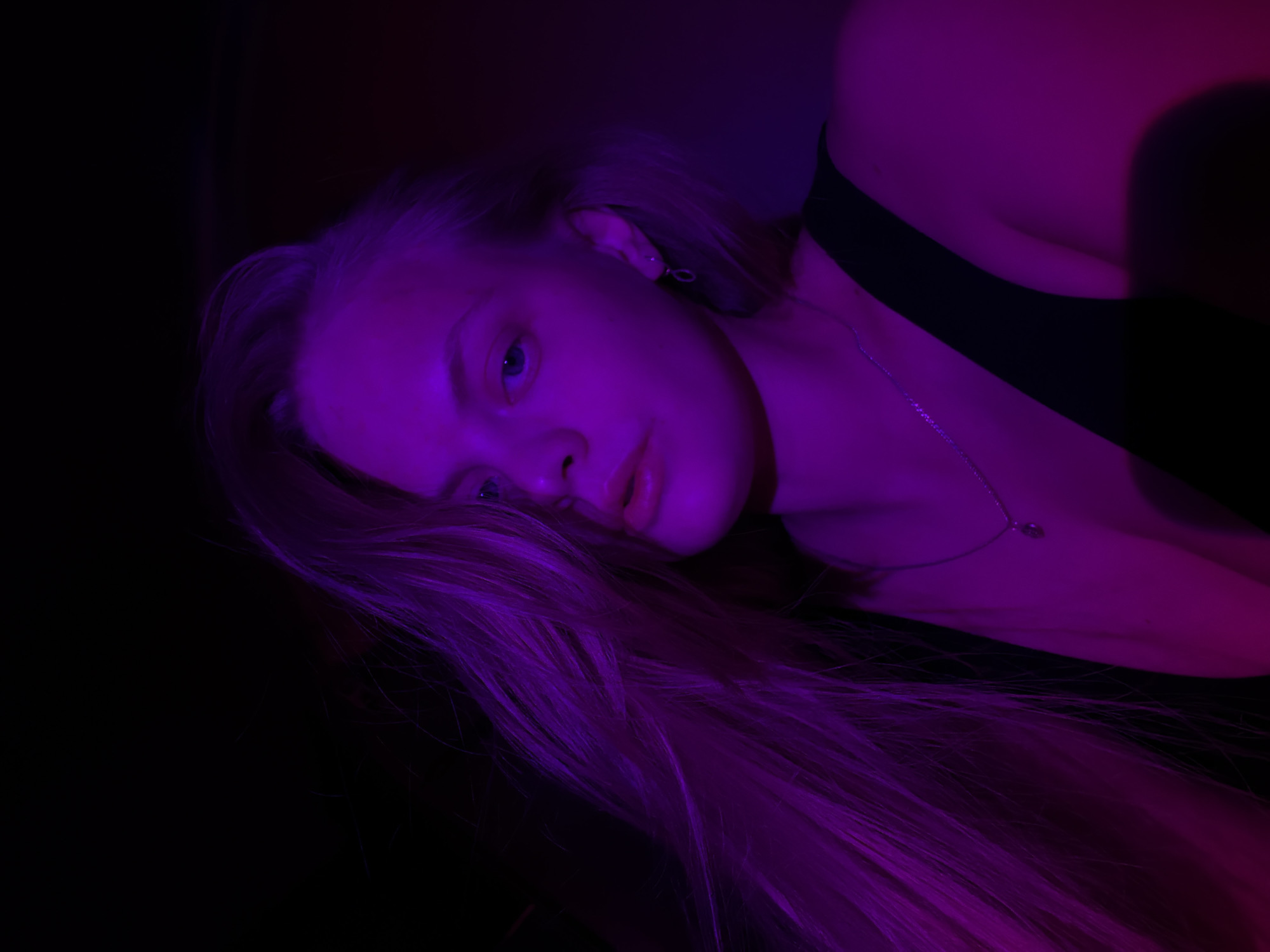 A woman with long blonde hair is lying down. She is wearing a black top and a silver necklace. The lighting is predominantly purple. The location is not discernible, and there is no indication the image was taken in Izhevsk, Russia.