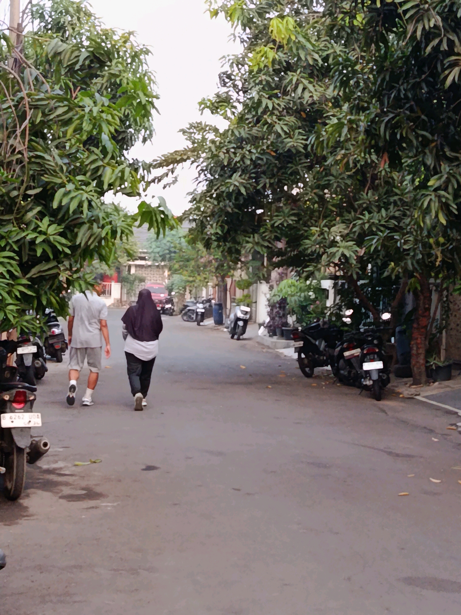 The image depicts a street scene in the city of Bekasi, Indonesia, likely in a residential area. Two individuals are walking away from the camera on the asphalt road. The person on the left is wearing a light grey short-sleeved shirt and light grey shorts, with white socks and dark shoes. The person on the right is wearing a dark headscarf and a light-colored top with dark pants and light-colored shoes.
The street is lined with trees that have dense green foliage, casting some shade on the road. On the sides of the street, motorcycles are parked. To the left of the road, there are several parked motorcycles, one of which is partially visible in the foreground with a license plate that reads "B 6262 UNA". On the right side of the street, more motorcycles are parked, and some potted plants are visible. In the background, houses with tiled roofs and walls are visible, along with a red vehicle and more parked motorcycles. The sky is overcast, suggesting it might be late afternoon or early morning. The overall impression is a quiet, everyday street scene.