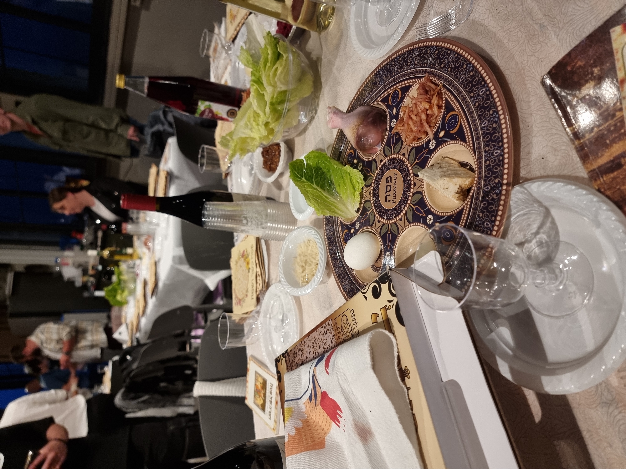 The image captures a Passover Seder table setting, likely indoors. Dominating the scene is a Seder plate with traditional symbolic foods arranged in designated sections. There's a shank bone, lettuce, charoset, matzah, a hard-boiled egg, and maror or horseradish paste visible. Additional Passover items like matzah boxes, bowls of shredded horseradish, lettuce, a bottle of wine, and glasses are also present. Several people are around the table, with a few in focus. The environment looks like a gathering in a home or event space. It is impossible to determine if the event took place in Tel-Aviv from the image, it only shows details of the Seder table and the gathering around it.