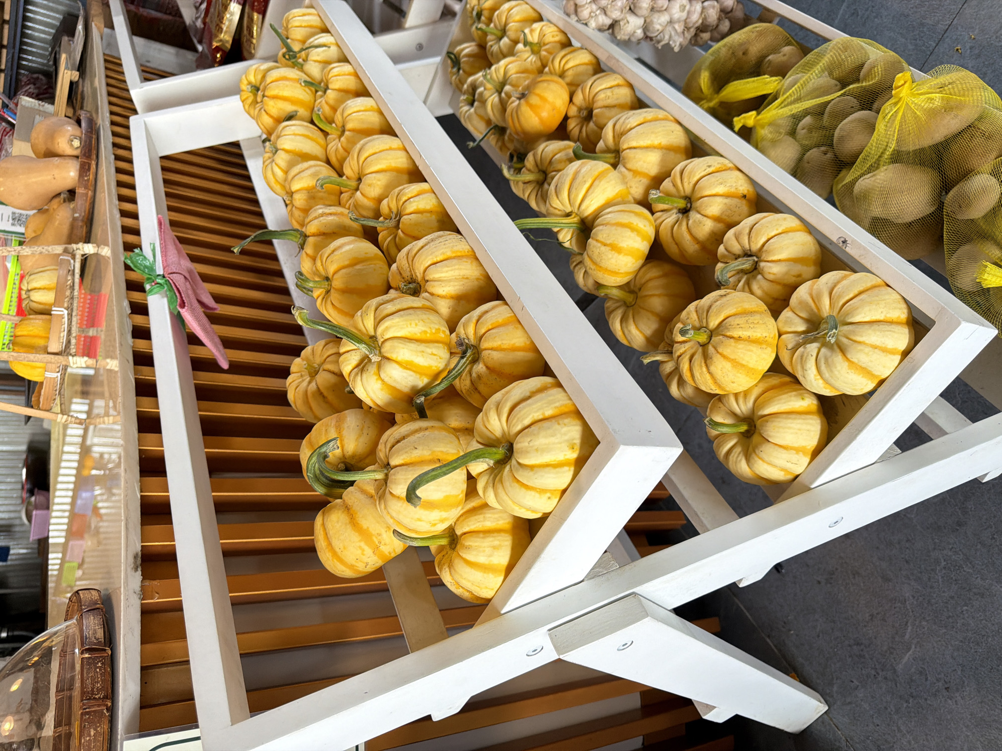 A display of mini striped pumpkins is shown on a white tiered stand in Da Lat, Vietnam. The pumpkins are orange and yellow striped with green stems. They are arranged in rows on three shelves. The bottom shelf also contains two yellow mesh bags filled with potatoes. In the background, there are other vegetables, including butternut squash and garlic, as well as some packaged goods. The scene appears to be at a market or a roadside stall.