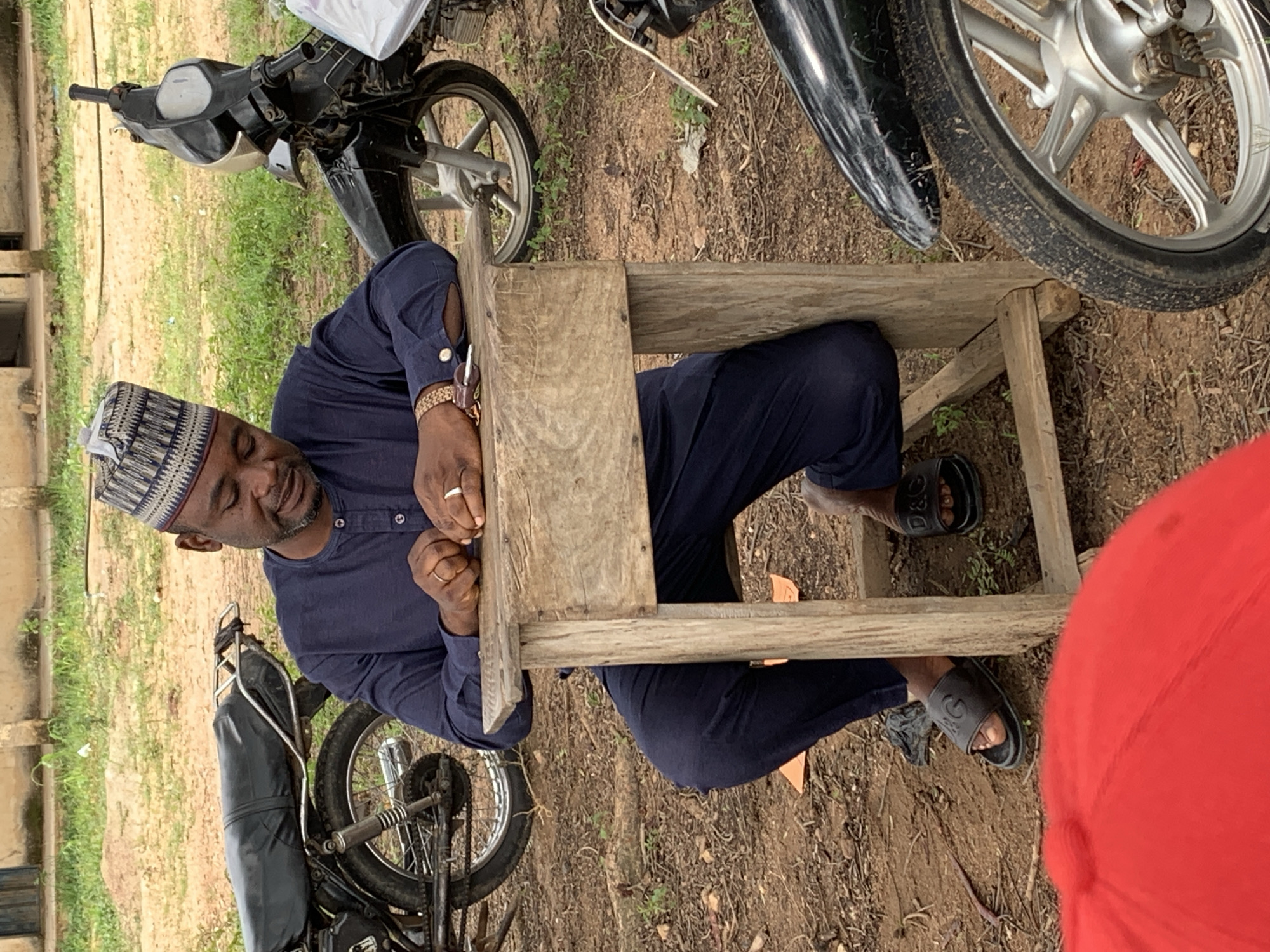 A man wearing a navy blue outfit and a patterned cap sits at a wooden desk outdoors. He is looking down at his hands, which are resting on the desk. He is wearing two rings on his fingers. Two motorcycles are visible in the background, one on the left and one on the right. The ground is covered in dirt and dry leaves. The scene appears to be in Tabu, Nigeria.