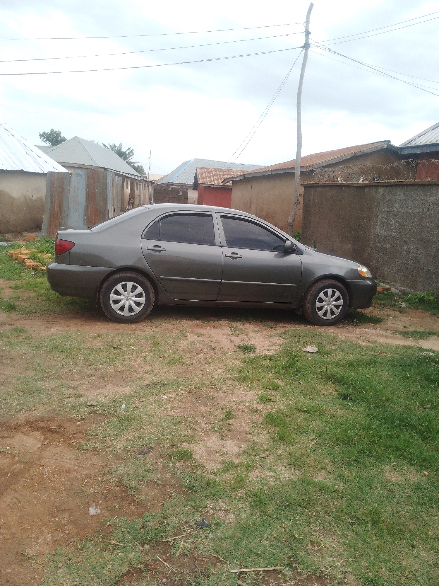 A dark gray sedan is parked on a dirt and grass lot. The car is facing right. Behind the car, there are several buildings with corrugated metal roofs and walls, as well as a concrete wall with barbed wire on top. A wooden utility pole with wires runs up the right side of the image. The sky is overcast.