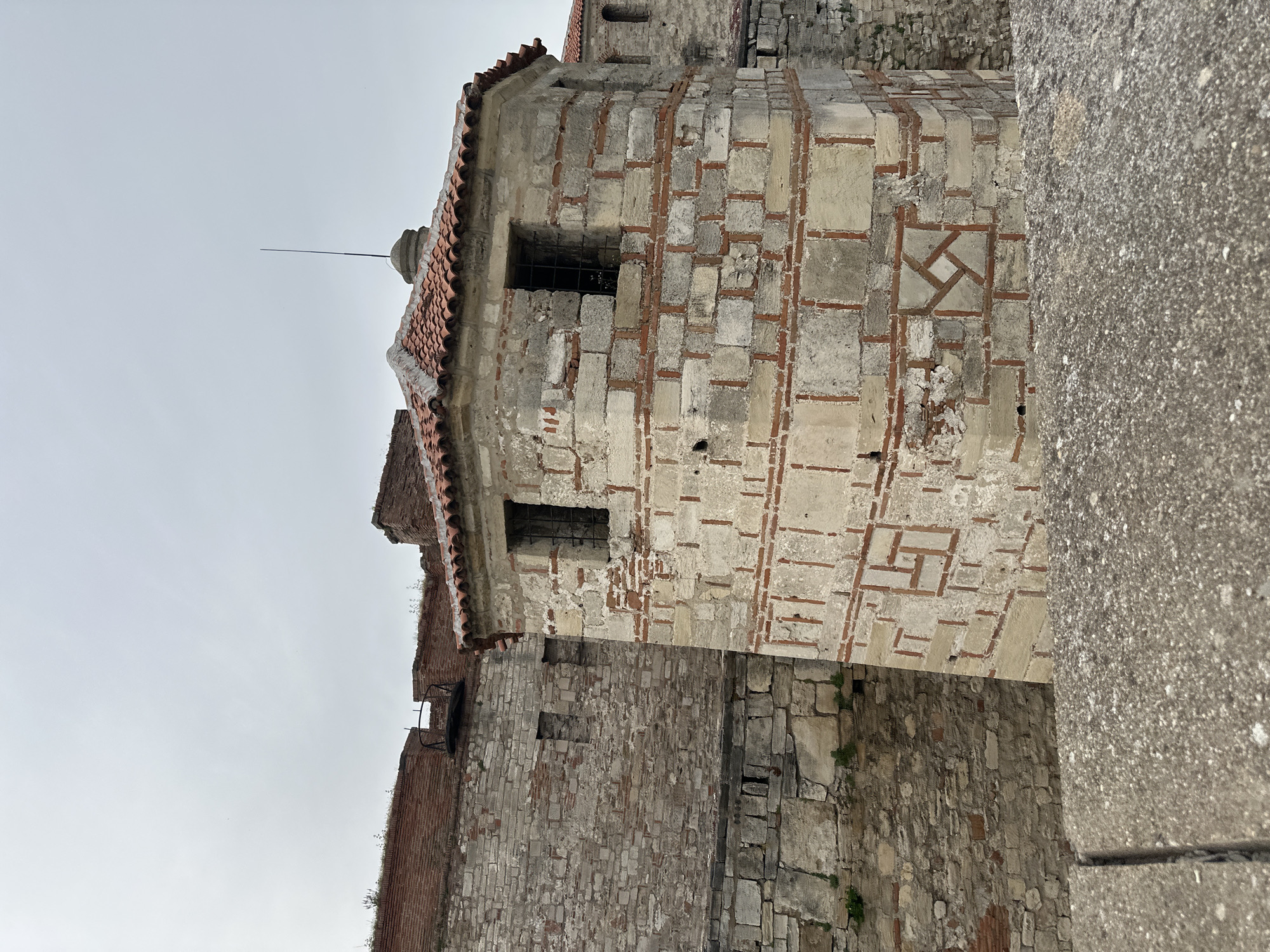 The image shows a section of the Baba Vida Fortress in Vidin, Bulgaria. The fortress walls are constructed of stone and brick, with a small, turret-like structure exhibiting a pattern of light-colored stone interspersed with red brick outlining the shapes. The structure has small windows with bars. The fortress is on a cloudy day, and a plain wall runs along the right.