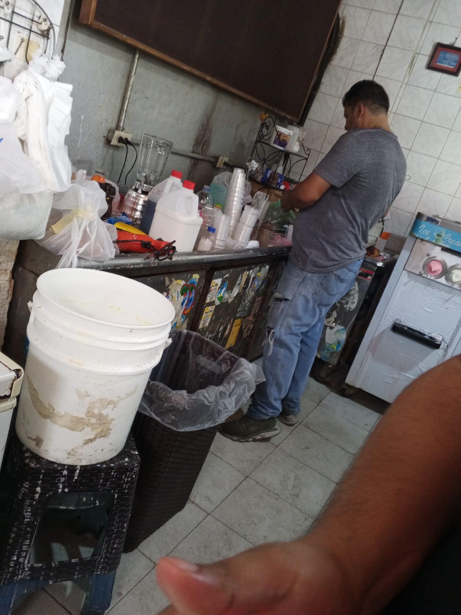 A slightly blurry, indoor scene depicts a man in a gray t-shirt and ripped blue jeans standing behind a counter, seemingly preparing food or drinks. He faces away from the camera, toward a cluttered workspace. The counter is covered with containers, bottles, and food preparation equipment. A large, white bucket sits atop a small, dark plastic stool in the foreground. A dark wastebasket is positioned next to the bucket. The background features tiled walls and a partial view of a machine labeled "Ice Cream". Part of a person's arm is in the foreground on the right side. The location is likely a small food preparation area in Tinaquillo, Venezuela.