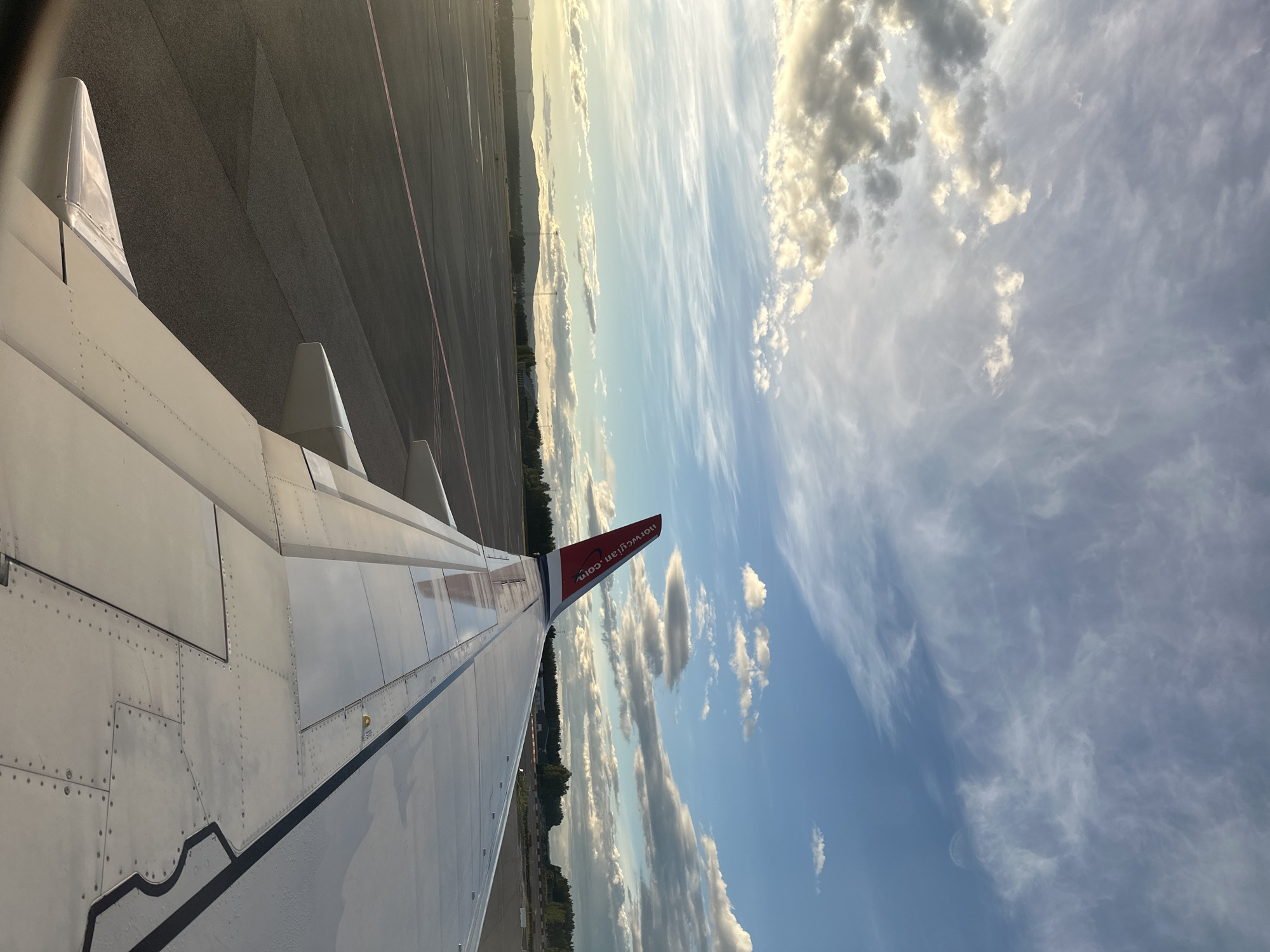 The image shows the wing of a Norwegian Air Shuttle aircraft on the tarmac at Sandefjord Airport, Torp (TRF), located in Sandefjord, Norway. The wing, which is white with rivets and panels, extends diagonally across the frame from the bottom right to the top left. The tail fin of the aircraft, featuring the Norwegian logo and text "norwegian.com," is visible at the top of the wing.
The tarmac is dark gray with a faint pink line running horizontally across it. In the background, a flat landscape of trees and distant hills under a dynamic sky filled with scattered clouds and streaks of blue sky is visible. The lighting suggests it is either early morning or late afternoon, with sunlight casting long shadows. There are no people or any other aircraft visible in the frame. The scene is static, depicting the aircraft on the ground at an airport.