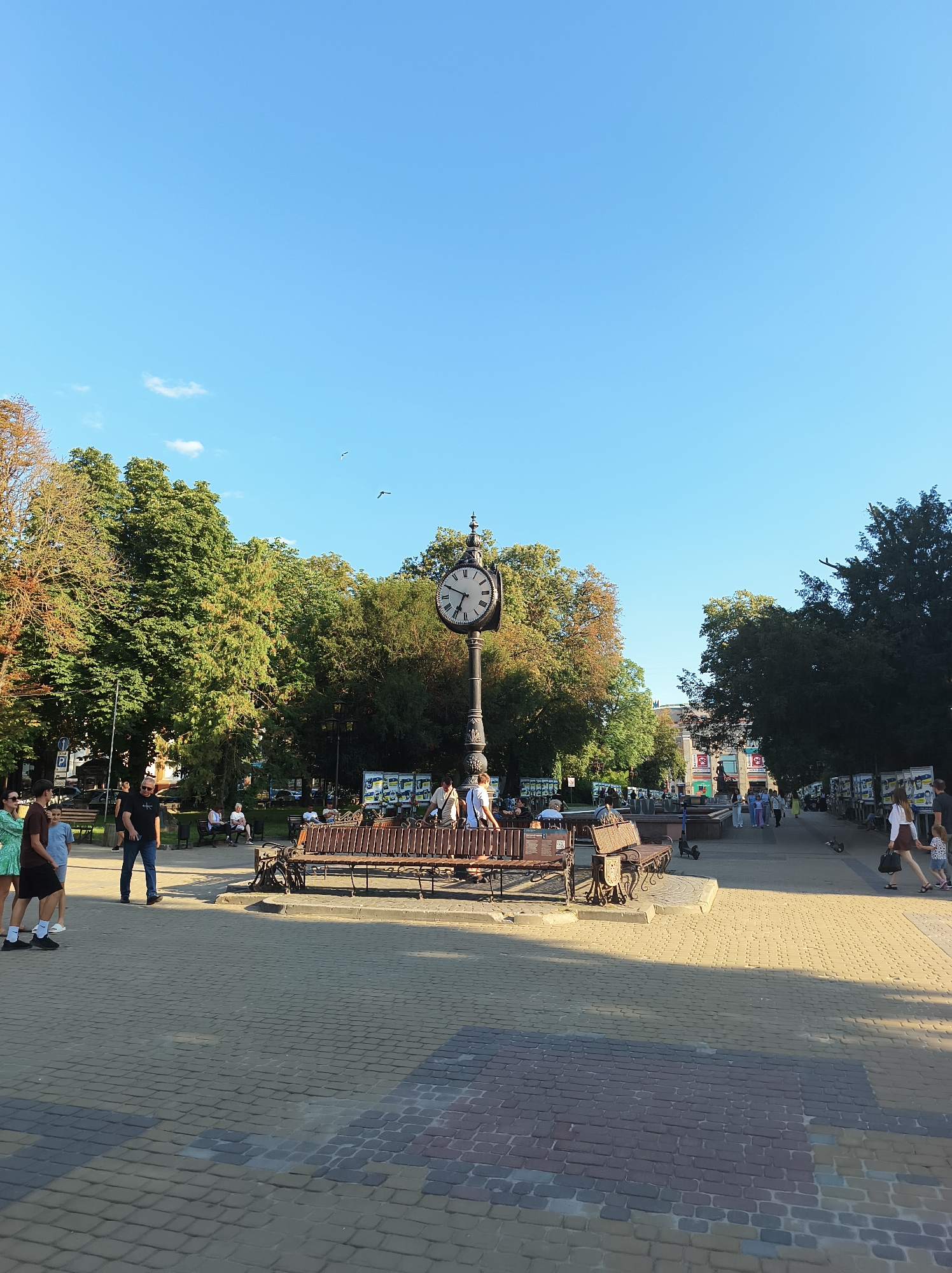 The image shows a public square in Ternopil, Ukraine, on a clear day. In the foreground, a paved walkway made of interlocking bricks leads into the scene. Several people are walking on the walkway, some sitting on park benches, and others standing around.
A prominent feature in the middle ground is a large, ornate clock tower with Roman numerals. Two park benches are positioned in front of the clock tower, one facing the camera and the other facing away. Behind the benches, trees with green foliage form a backdrop.
Further in the background, a fountain is visible, with more people gathered around it. A building with a red facade and posters on its exterior is also visible in the distance. The sky above is a clear, bright blue with a few small clouds and two birds in flight.
The overall scene depicts a lively public space with elements of nature and urban architecture. The light suggests it is late afternoon or early evening, with the sun casting long shadows across the paved area.