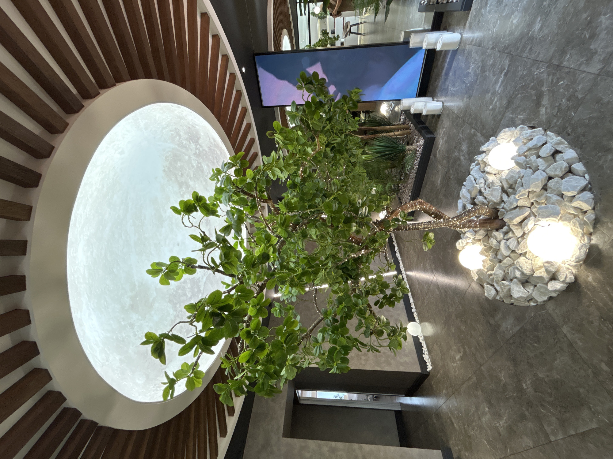 The image depicts an indoor scene with a central, artificial tree featuring green leaves and decorative lights on its branches. The tree is planted in a circular bed filled with white rocks and illuminated by several globe-shaped lights at its base. Above the tree, a large, circular ceiling light fixture resembles the moon, surrounded by wooden beams. To the right of the tree, a tall, vertical digital screen displays a blue and purple abstract image. The floor is tiled with large, gray, marbled tiles. In the background, a doorway leads to another room, and more plants and architectural features are visible. The location is identified as Votkinsk, Russia. There are no people clearly visible in the image.