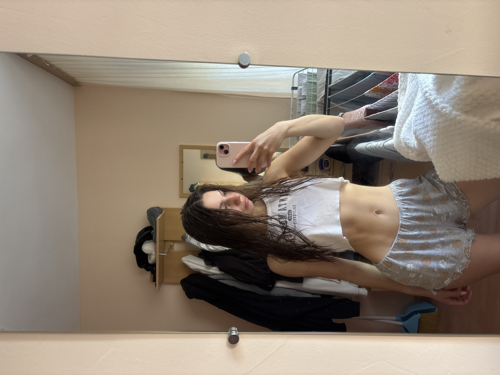 The image captures a mirror selfie taken by a young woman, with the entire picture rotated 90 degrees clockwise. The woman stands before a large rectangular mirror, holding a pink-cased smartphone to take the photo. Her long, dark hair appears wet, and she looks directly at the camera with a neutral expression. She wears a white crop top with mirrored text that reads "WHAT'S UP" and light grey shorts patterned with cartoon bears and milk bottles.
The setting is an indoor room with light beige-colored walls and a lighter ceiling. In the reflection, to the left of the woman, a wooden coat rack or shelf holds several items, including dark and light clothing and possibly a white fluffy item. To the right, a multi-tiered clothes drying rack is visible, holding various garments, including a large white fluffy towel or blanket hanging over the side. Below the woman's torso, reflected on the floor, is a blue broom and dustpan set. A small grey circular object is adhered to the wall above the mirror, and another silver hook is visible lower down. The overall scene suggests a casual, domestic environment, possibly a bedroom or utility area.