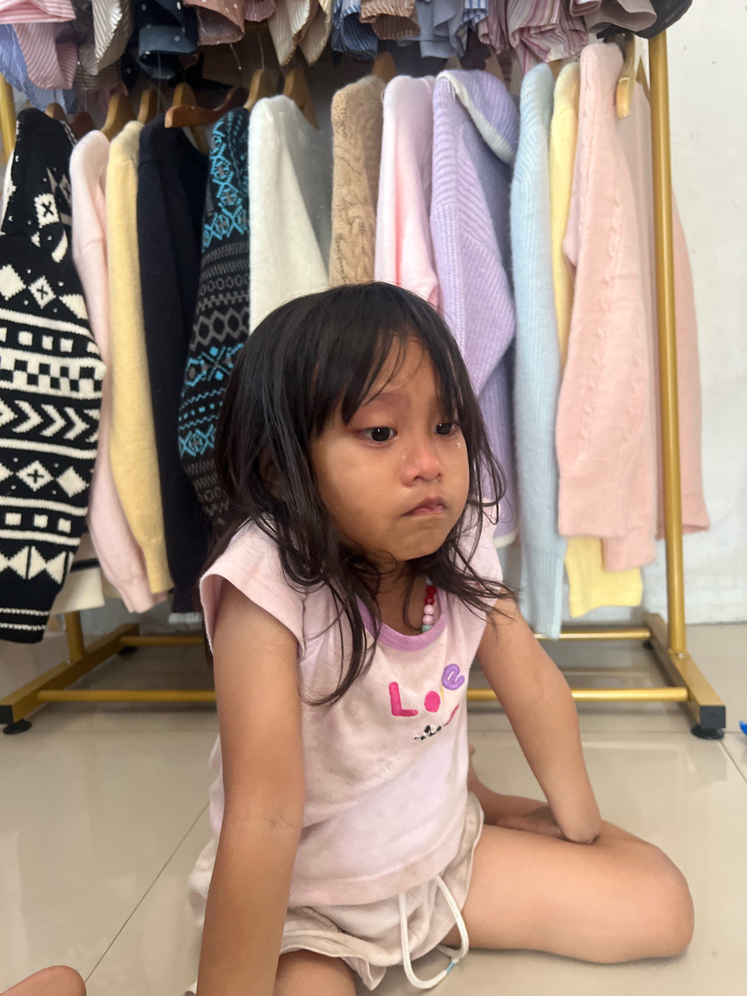 A young girl with dark hair is sitting on the floor in front of a clothing rack. She is wearing a light pink t-shirt with "LO" and a smiley face on it, and light pink shorts. Her face is turned slightly to the side, and she appears to be crying, with tears visible on her cheeks and nose. The clothing rack behind her is filled with various colored sweaters and clothing items. The setting appears to be indoors, possibly a bedroom or a store, with a tiled floor. The overall mood conveyed is one of sadness or distress due to the girl's expression. The location is specified as Kecamatan Pati, Indonesia.