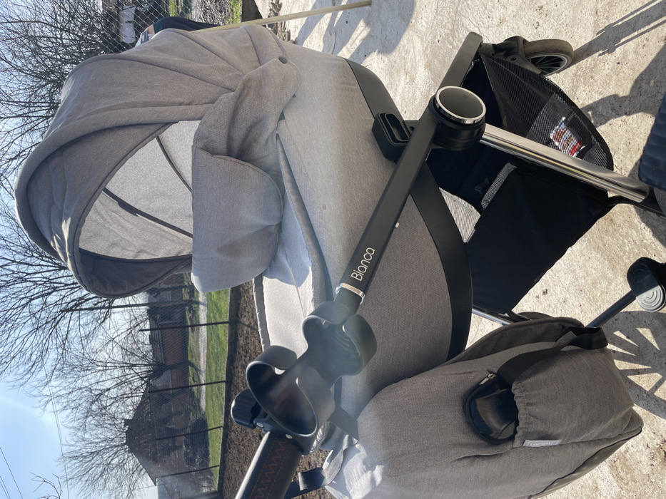 A grey 'Blanco' brand baby stroller, configured with a carrycot, is stationary on a light-colored concrete surface. The stroller features a fully extended grey canopy and matching bassinet. A black cup holder is attached to the main handle, which has a textured brown grip. A grey utility bag, containing a black object in an exterior pocket, hangs from the left side of the stroller frame. A black mesh storage basket is located underneath the bassinet. The background reveals a disturbed garden plot with dark soil, bordered by a wire fence. Beyond the fence, bare deciduous trees and a building with a red roof are visible under a clear, bright blue sky. No individuals are present. The setting is in the city of Tarnava, Bulgaria.