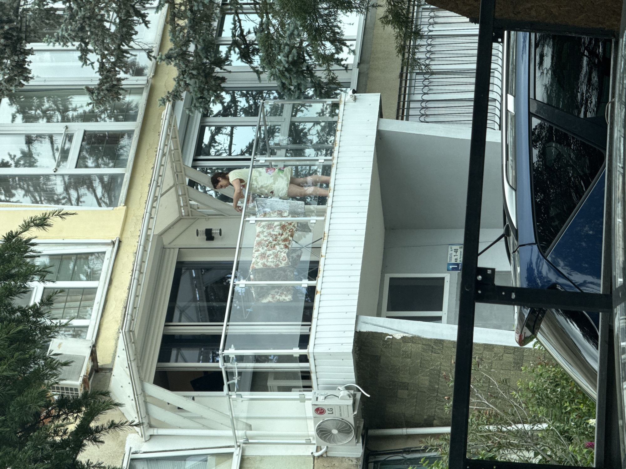 A daytime, medium shot captures a building, possibly an apartment complex, in an urban environment in Unknown, Unknown. The architecture features windows, balconies, and an air conditioning unit. A person is visible on one of the balconies, possibly interacting with household items or the balcony structure itself. A parked blue car is partially visible on the right side, with parts of a metal structure possibly obstructing the view. Vegetation is visible in the background and near the building. A street number sign is visible on the wall of the building: 1 кВ. 1-48.