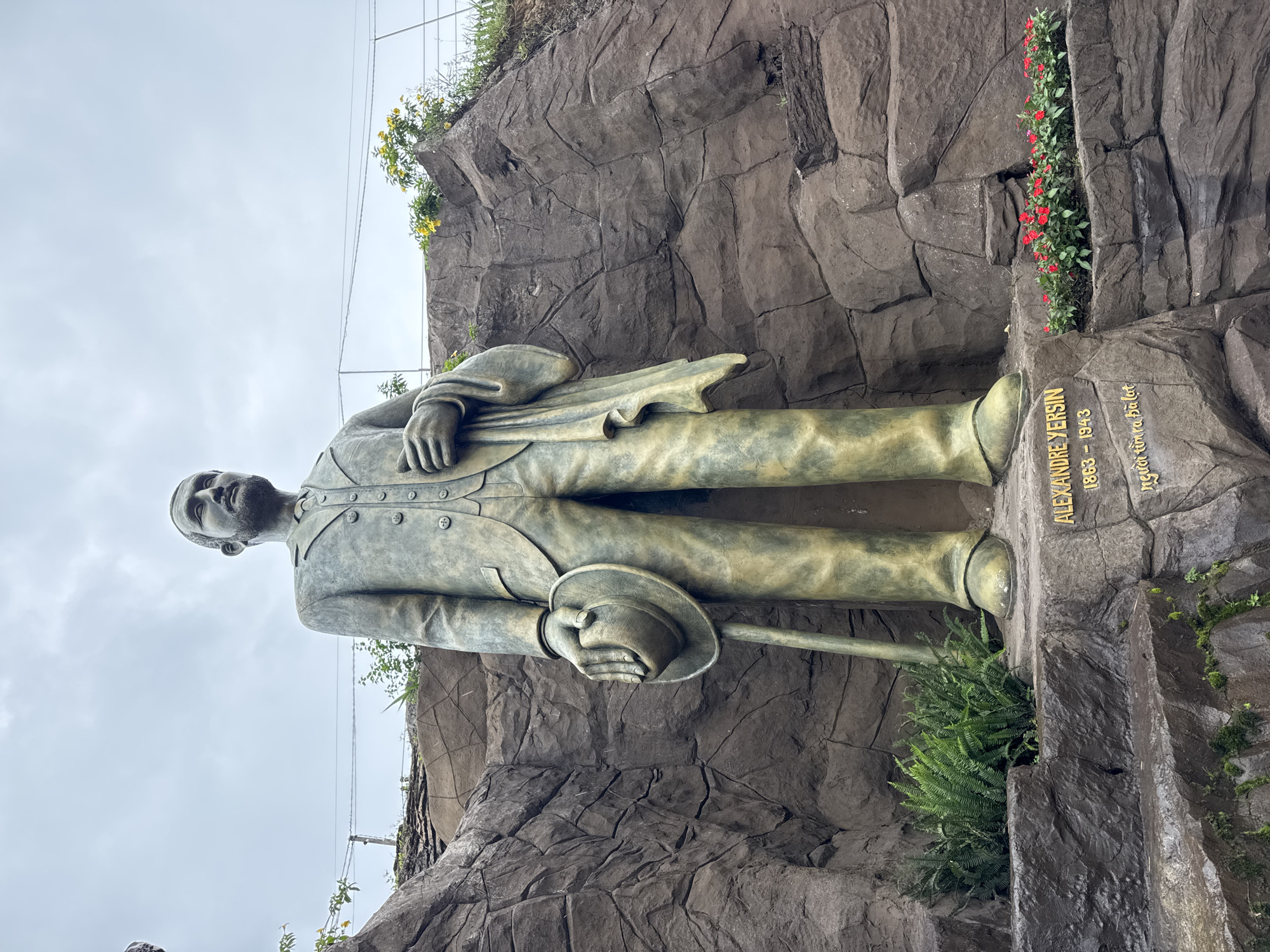 The image shows a bronze statue of Alexandre Yersin, a Swiss-French bacteriologist who founded the Pasteur Institutes in Nha Trang and Dalat, Vietnam. The statue is located in Dalat, Vietnam, and depicts Yersin in a suit and tie, holding a hat in his left hand and a cane in his right. He is standing in front of a rocky cliff face, with lush green ferns and red flowers planted at his feet. The sky is overcast and the overall mood of the image is somber and contemplative. The inscription on the base of the statue reads "ALEXANDRE YERSIN 1863 - 1943" and "người tìm ra Đà Lạt" (the person who discovered Dalat).