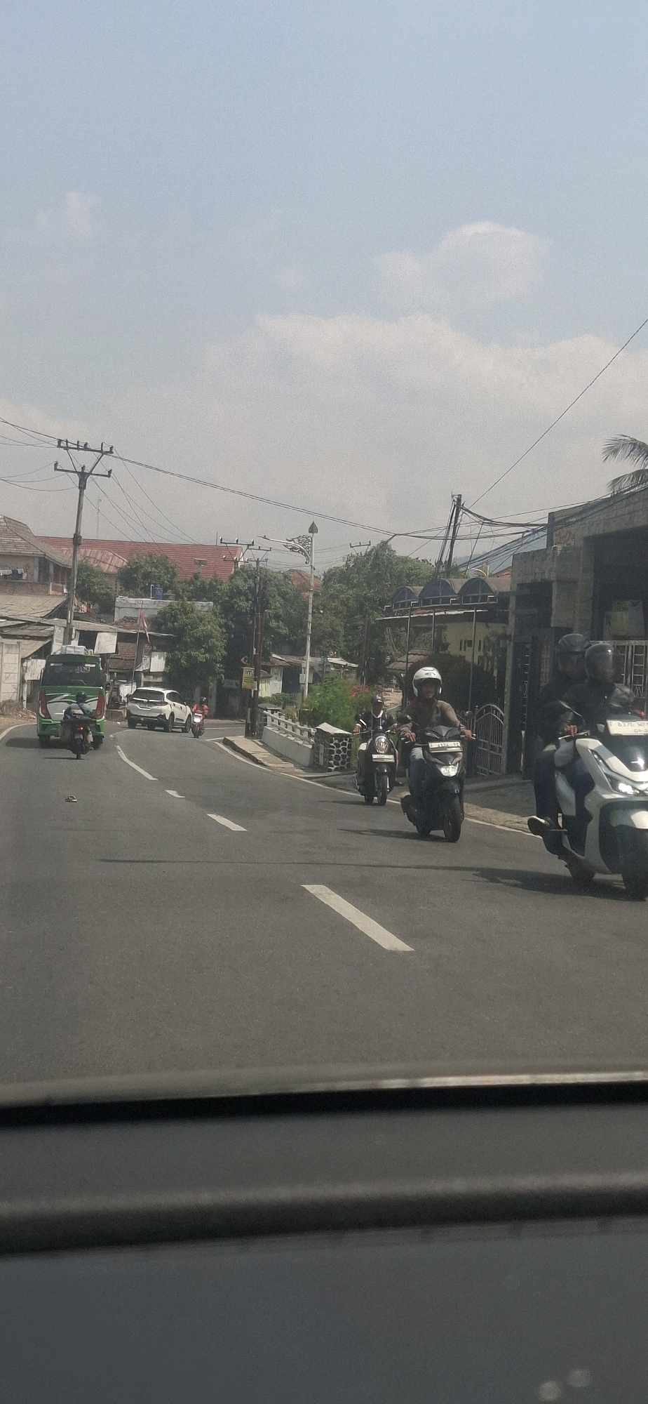 This image captures a street scene in Sukasari, Indonesia, during daylight. The asphalt road is marked with white lane dividers, indicating multiple lanes of traffic. Several vehicles are present, including a green bus, a white car, and multiple motorbikes. The bus is on the left side of the road, and the white car is further up the road. Motorbikes are scattered throughout the scene, with riders wearing helmets.
On the left side of the road, there are buildings with red tiled roofs, some appearing to be houses or commercial establishments. Utility poles with visible electrical wires are prominent along the roadside. Trees with lush green foliage are interspersed between the buildings, suggesting a moderately vegetated urban area.
On the right side of the road, a building with a dark facade and possibly a metal gate is visible. A concrete barrier with a decorative pattern lines the edge of the road. The sky is bright and clear with some scattered clouds. The overall impression is that of a typical street in a populated area of Indonesia, with a mix of residential and commercial structures and active traffic.