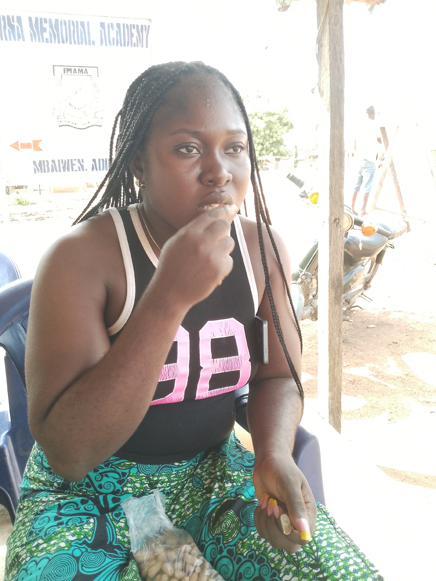 In Ichol, Nigeria, a young woman with braided hair is seated outdoors, her gaze directed downwards as she brings food to her mouth. She wears a black tank top with pink "98" graphics and a vibrant green and teal patterned skirt. Her hands are adorned with long, yellow and pink manicured nails and rings. A clear plastic bag filled with nuts rests beside her on her lap, near her skirt. The background reveals a wooden post, a motorcycle, and a sign that reads "IMAMA" and "MBAWLEN. ADI". A distant figure stands near a wooden structure. The lighting suggests it is daytime.