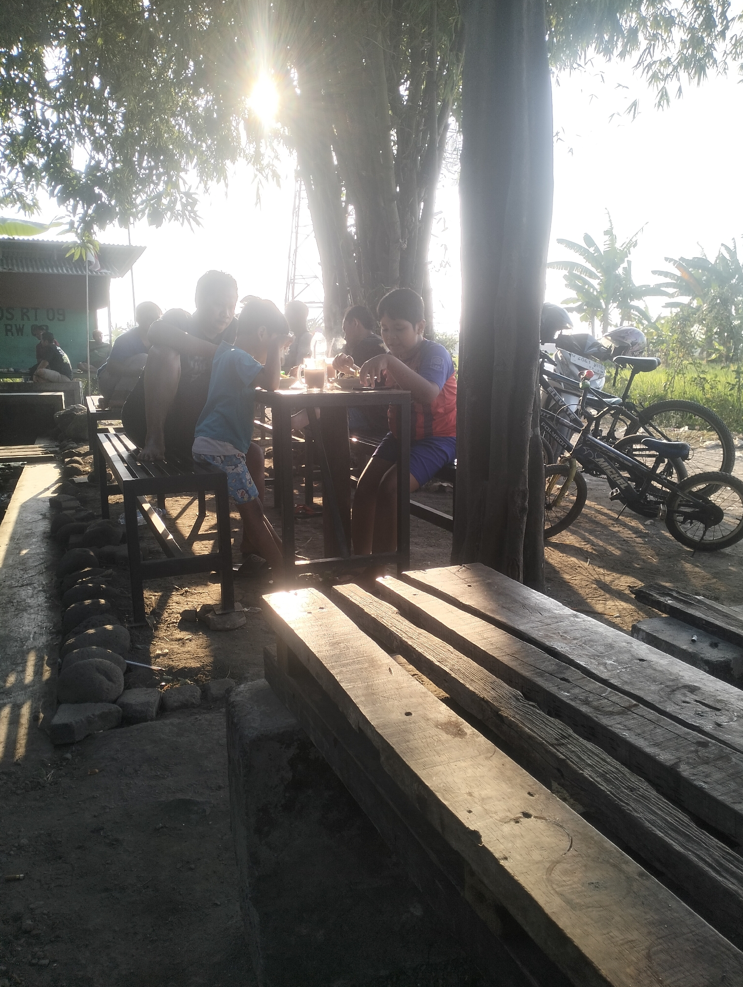 In Sambungrejo, Indonesia, a group of people, including several children, are gathered outdoors at dusk. They are seated around a table with drinks, possibly a local eatery. The scene is illuminated by the setting sun, casting long shadows and creating a warm, golden light. The environment appears rural, with trees and vegetation in the background, and a simple structure that may be a shop or restaurant. Several bicycles are parked nearby, suggesting a mode of transport common in the area. The overall impression is one of a casual gathering in a peaceful, natural setting.