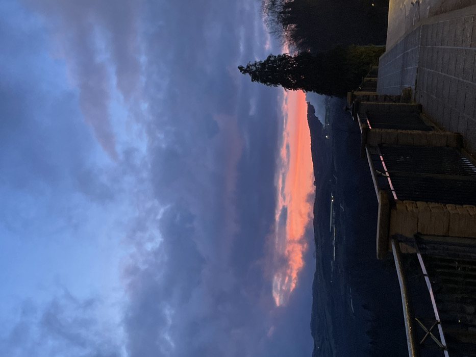 The image captures a serene twilight scene over a mountainous landscape, likely in Santa Fiora, Italy. The time of day is clearly sunset or dusk, indicated by the dramatic and colorful sky. A prominent horizontal band of vibrant orange and pink clouds stretches across the middle of the frame, illuminated by the last rays of the sun. Above and below this fiery display, the sky transitions into deeper blue and purple hues, filled with various grey cloud formations that add texture and depth.
In the foreground, on the right side, a paved pathway or street slopes downwards, bordered by stone-like structures and dark metal railings. A tall, dark green coniferous tree, possibly a cypress, stands silhouetted against the bright sky, adding a vertical element to the composition. Beyond the immediate foreground, the landscape unfolds into a vast, dark valley with the silhouettes of distant hills or mountains. A few faint lights twinkle in the far distance, suggesting remote settlements or buildings. No people or specific activities are visible, emphasizing the quiet, natural beauty of the moment. There is no visible text in the image. The striking contrast between the warm sunset colors and the cooler, darker tones of the landscape creates a captivating atmosphere.