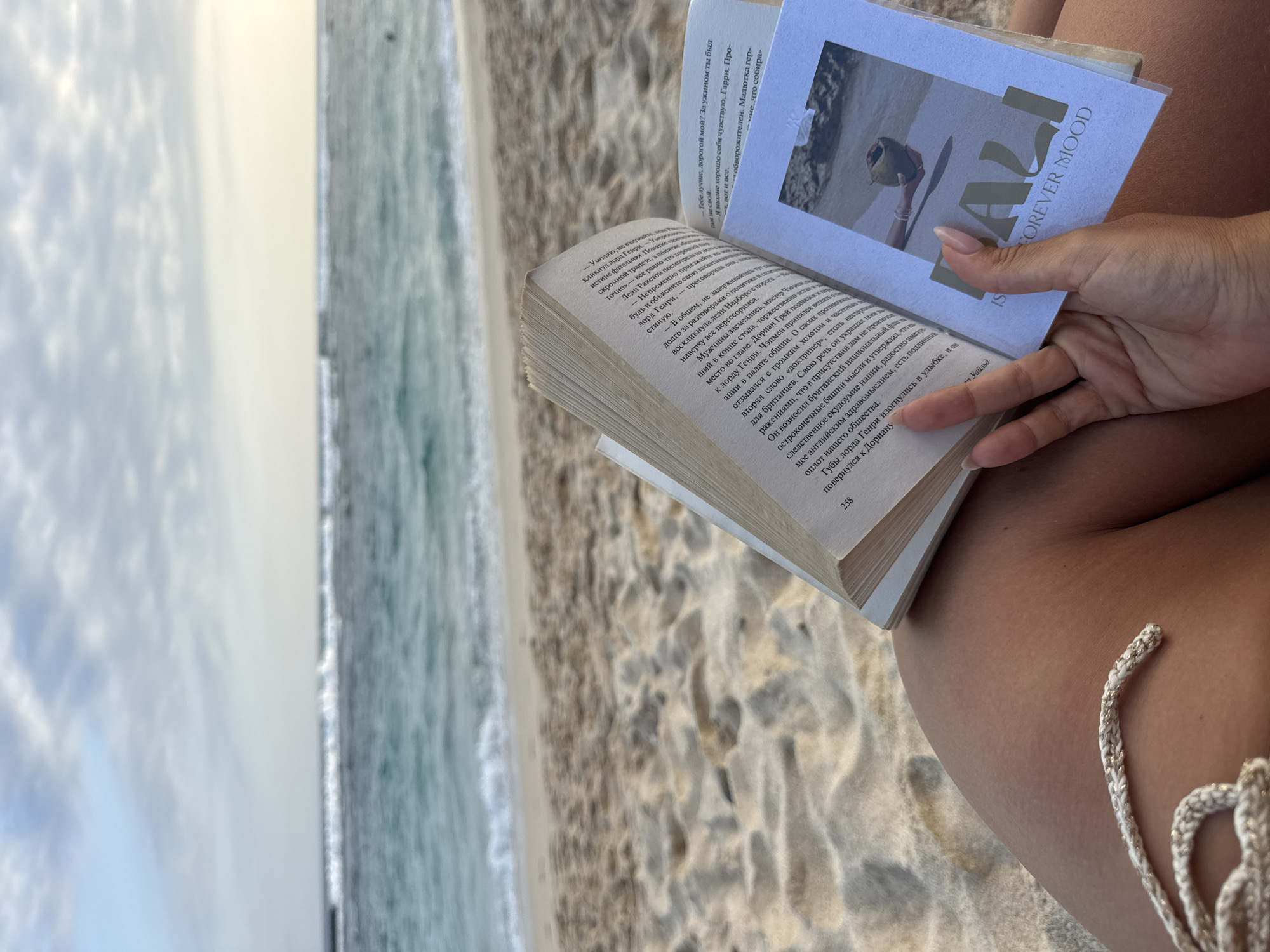 A close-up shot shows a woman's hand holding an open book on a sandy beach. The book is positioned over her lap, which is covered by a light-colored string bikini bottom. The pages of the book display text and an image of a coconut on a beach. The background features the ocean with gentle waves breaking and a hazy sky. The location is identified as Kuta Selatan, Indonesia, suggesting a beach setting within that region. The scene appears to capture a moment of relaxation and reading by the sea.