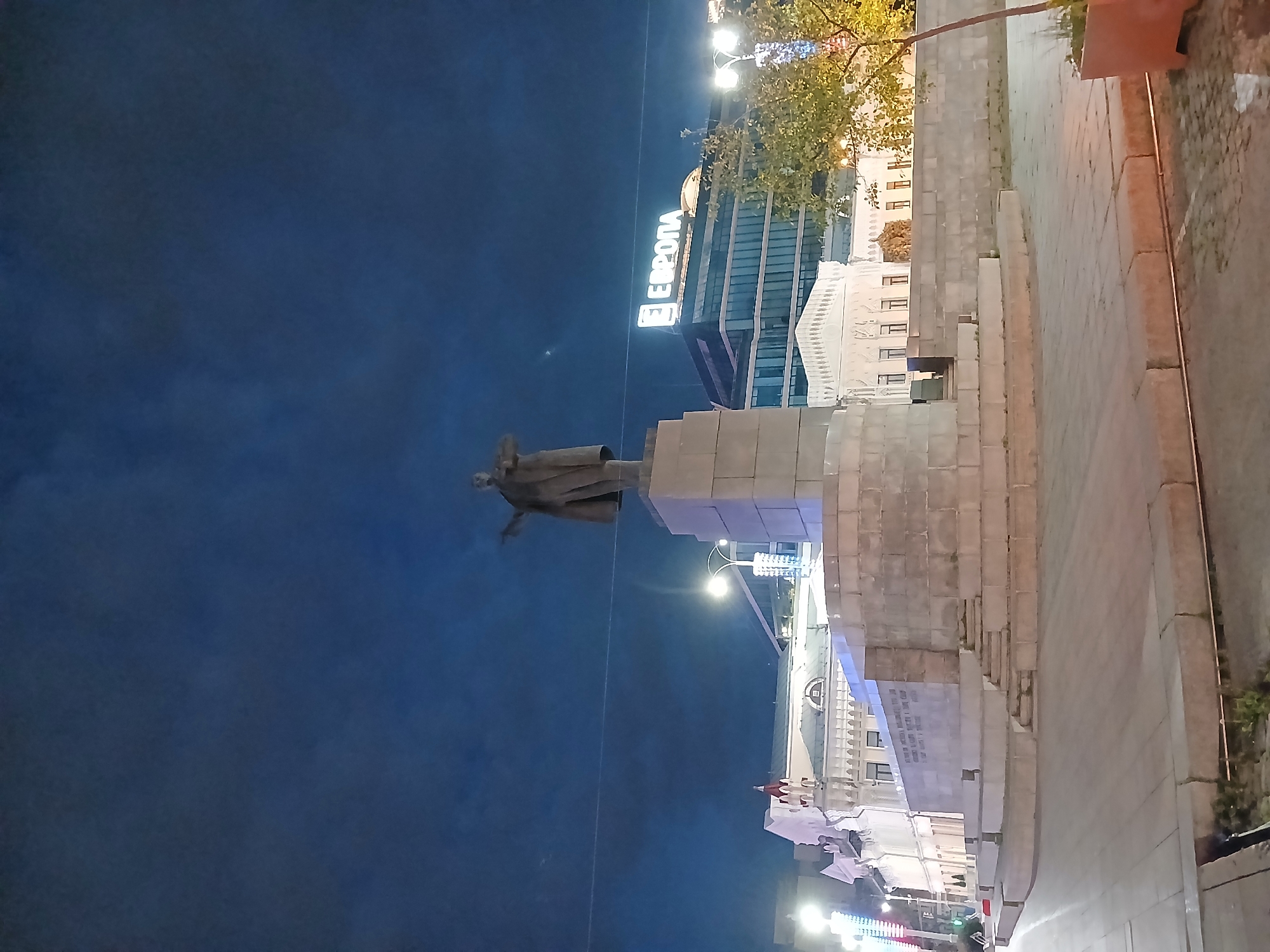 This is a night-time, eye-level, full shot of the monument to Vladimir Lenin located in Yekaterinburg, Russia. The monument stands on a large, multi-tiered stone pedestal. Lenin is depicted in a full-body pose, wearing a coat and hat, with his right arm outstretched, pointing forward. Behind the monument, a modern building with illuminated signage spelling "ЕВРОПА" (Europe) is visible. To the left of the monument, there are other buildings with illuminated facades and a street light. The ground is paved with stone slabs. A small tree with yellowing leaves is situated in the foreground to the right, in a planter. The sky is dark blue, indicating night.