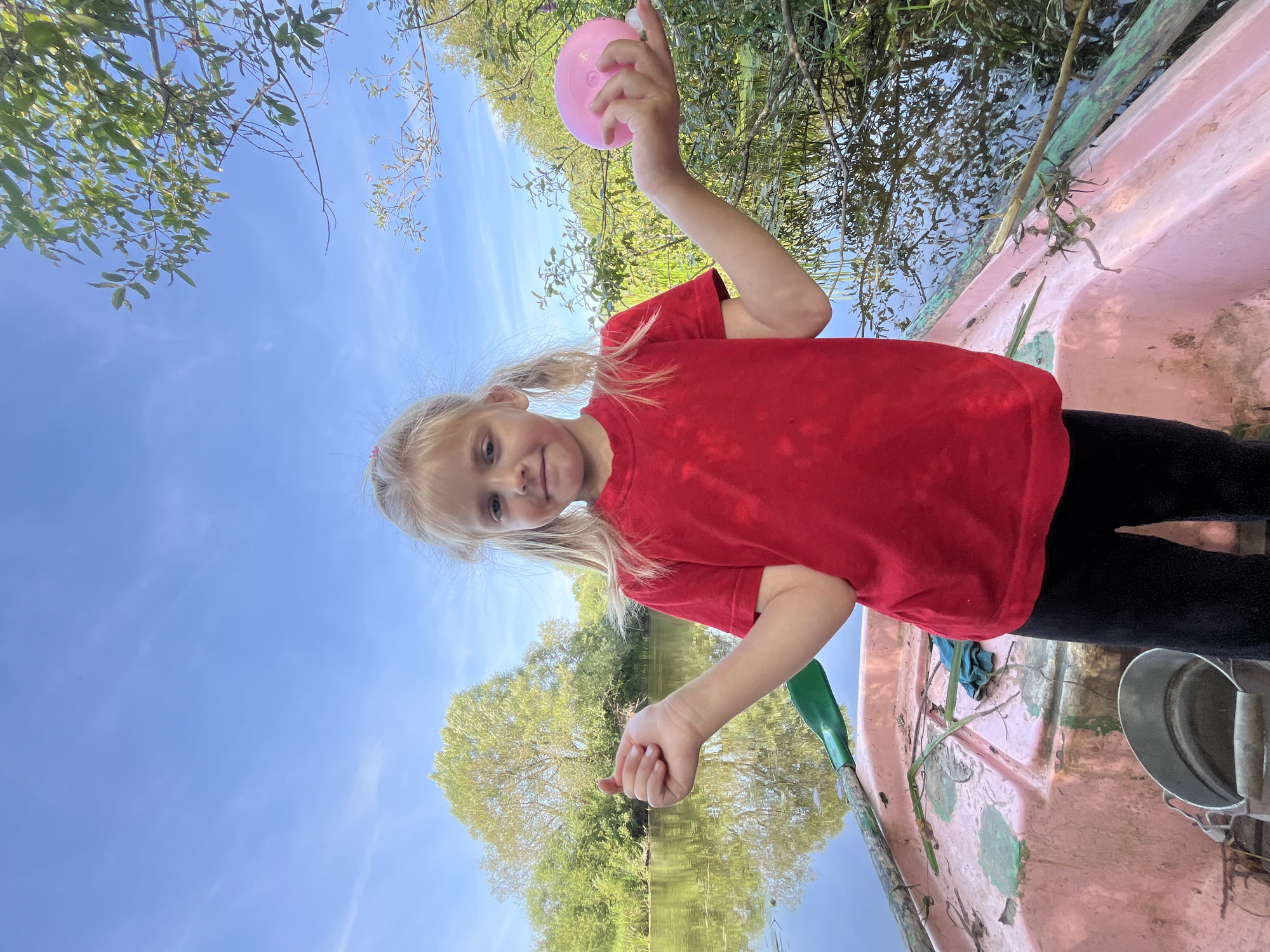 A young girl with blonde pigtails and blue eyes, wearing a red t-shirt and black pants, stands in a pink rowboat on a calm body of water. She holds a pink, round object in her right hand and gestures with her left hand. Green foliage and trees line the distant shore under a clear blue sky. The boat contains a green oar and a metal bucket. The location is Sharkawshchyna, Belarus.