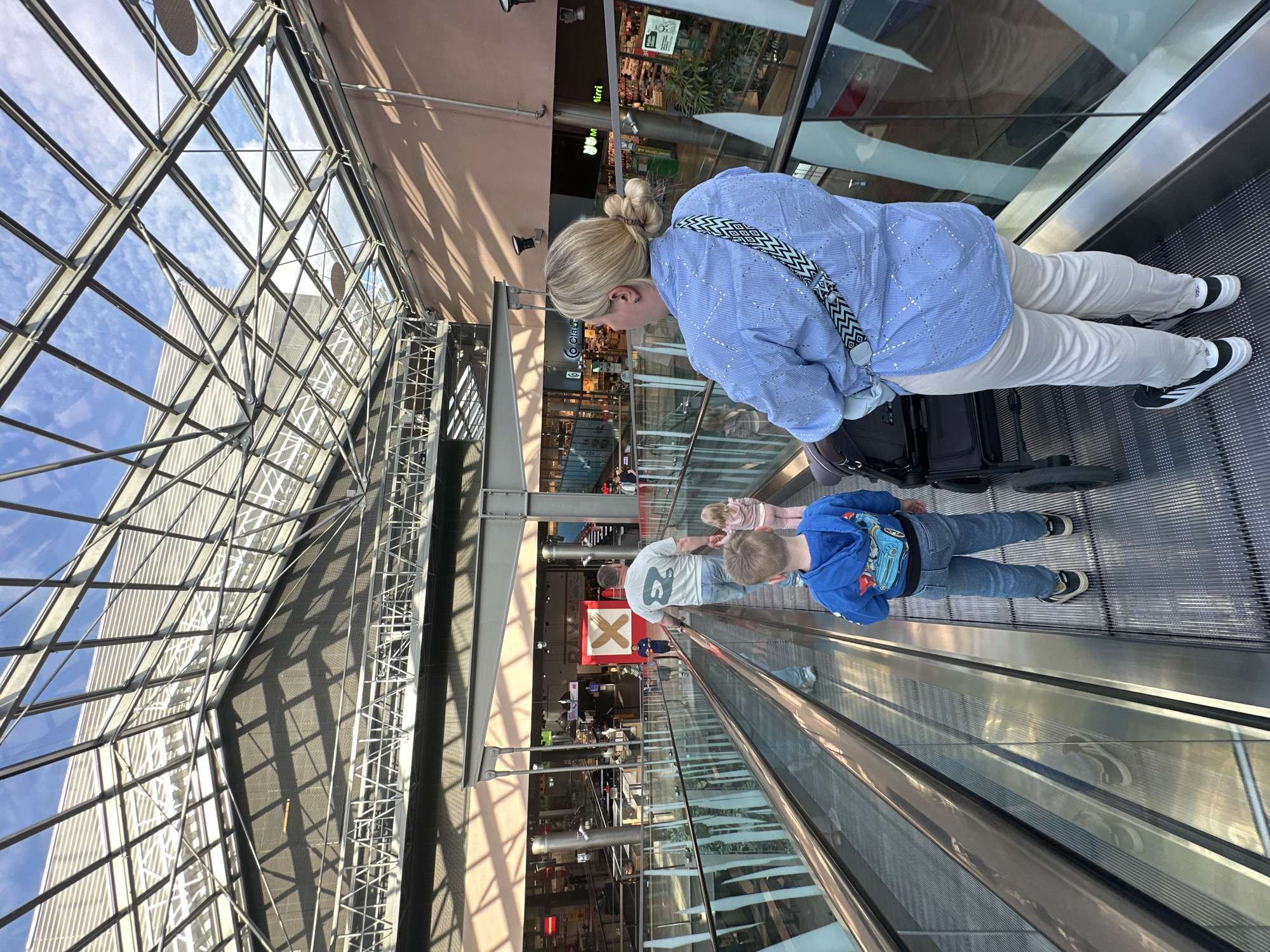 This media file shows a family on an escalator in what appears to be a modern airport or large indoor shopping mall. The scene is brightly lit, suggesting it is daytime. The architecture features a large glass ceiling with visible metal supports, allowing natural light to flood the space.
The primary subjects are a woman, a man, and two children. The woman is at the front, facing away from the camera, wearing a light blue patterned shirt and light-colored pants, and carrying a patterned bag. She is on an escalator going up. Behind her, a man in a grey t-shirt and blue jeans is also on the escalator, holding onto the handrail. Two children are near him. A younger boy in a blue hooded sweatshirt with a car graphic is in the foreground, also on the escalator. Another child, a girl in a pink outfit, is visible behind the man. The family appears to be traveling or shopping together.
The escalator itself is a prominent object, made of metal with ribbed steps. Luggage is visible near the woman. In the background, shops with various displays and signs are visible, further indicating a commercial or travel hub. One sign prominently features a red background with a white symbol of crossed cutlery, suggesting a food establishment.
The overall impression is one of a busy, functional space filled with natural light. The people seem to be engaged in the everyday activity of using an escalator in a public place. No specific emotions are clearly discernible from the individuals' postures or expressions, but the general atmosphere seems neutral.