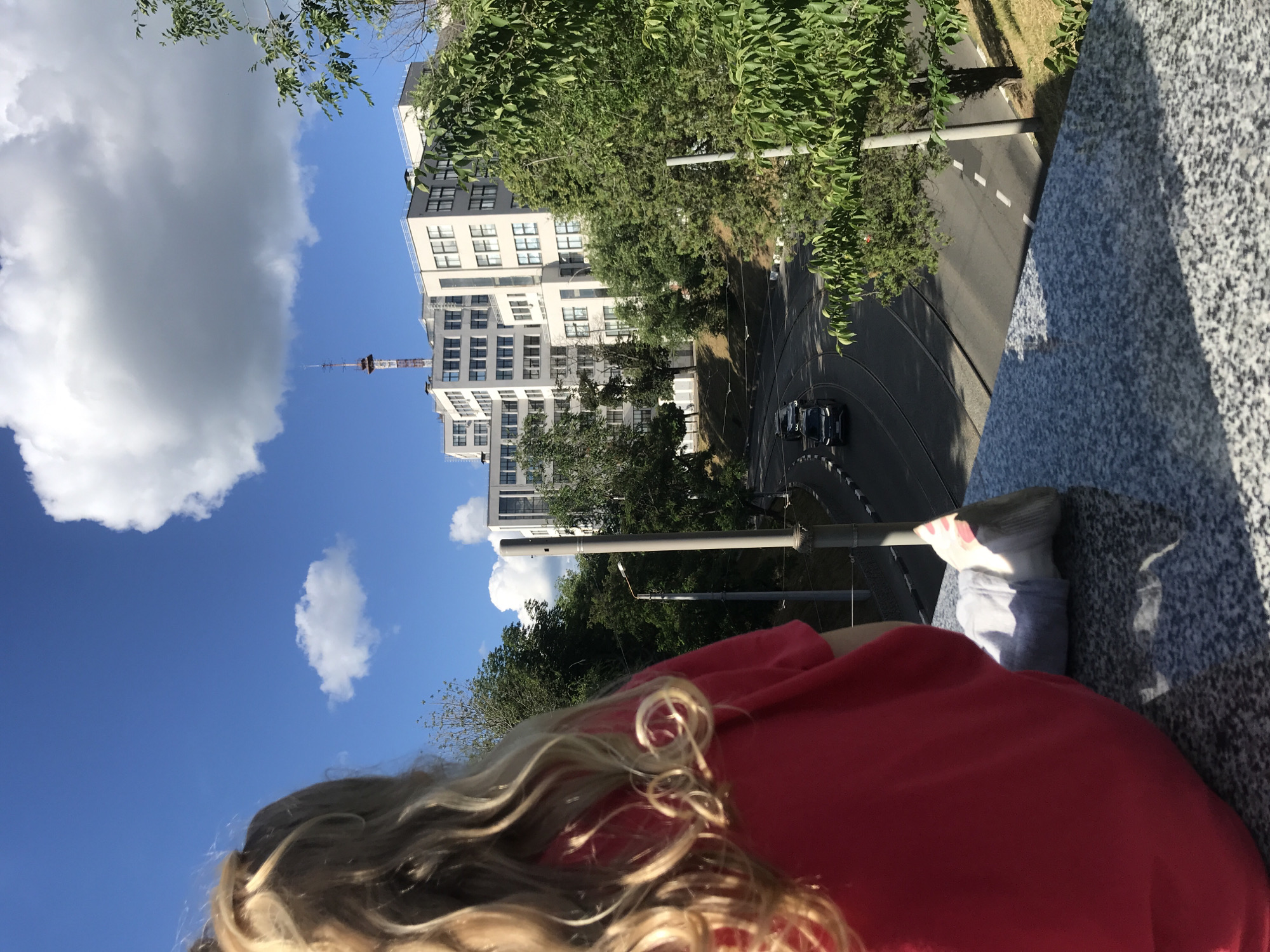 The image shows a blonde-haired child in a red shirt sitting on a granite ledge, looking out at a city scene. In the background, a large, white, multi-story building with many windows stands against a bright blue sky with white clouds. A radio tower is visible atop the building. Below the building, a road with tram tracks curves to the left, with two dark-colored cars driving on it. Lush green trees and vegetation line the road and surround the building. The scene appears to be set in Kharkiv, Ukraine, given the architectural style and the presence of tram tracks, a common feature in Ukrainian cities.