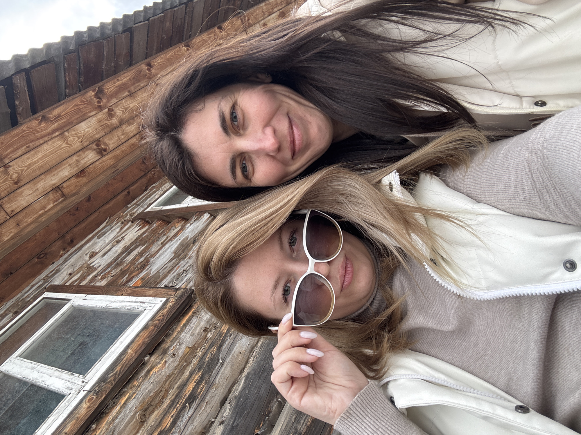 Two women pose for a selfie in front of a weathered wooden structure with white-framed windows. One woman has long brown hair and is wearing a cream-colored jacket. The other woman has blonde hair and is wearing sunglasses, a tan turtleneck, and a cream-colored jacket. The photo appears to be taken in Amaneevo, Russia.