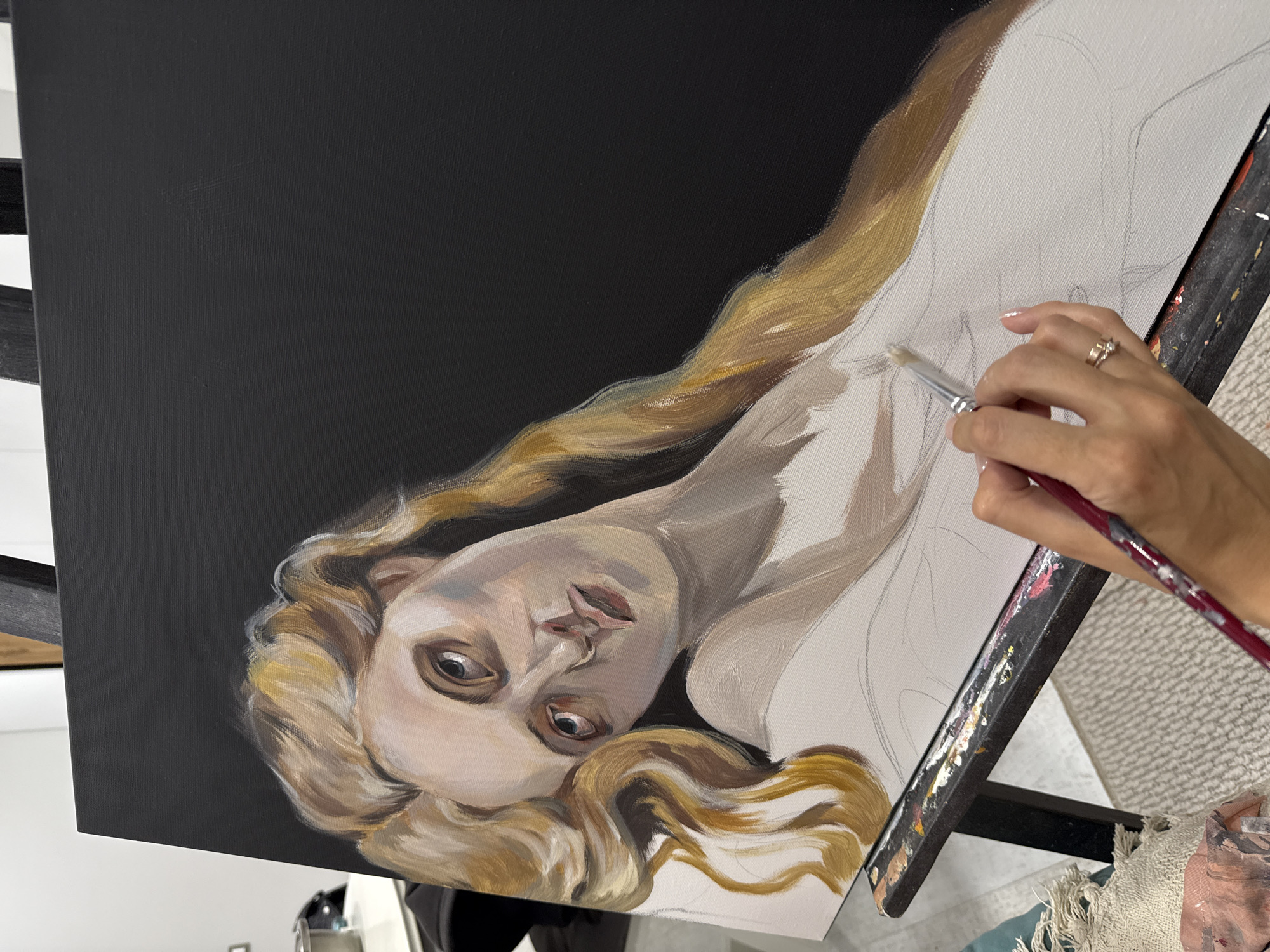 The image shows a close-up perspective of an artist's hand painting on a canvas in Krasnoyarsk, Russia. The main subject is an oil painting in progress, featuring a female figure with fair skin and long, wavy blonde hair. Her head is tilted, and her wide, intense eyes gaze upwards, conveying a sense of surprise or deep contemplation. Her lips are slightly parted.
An artist's right hand, adorned with a multi-banded ring and manicured nails, holds a paintbrush. The brush is actively applying paint to the canvas, specifically on the figure's upper body or clothing, which is still in its initial sketched form on the unpainted canvas. The face and hair of the figure are more developed, showcasing visible brushstrokes and nuanced tones.
The painting features a solid black background, providing a strong contrast to the lighter skin and hair of the figure. The canvas is supported by a dark easel, which is splattered with various colors of paint, indicating frequent use. A light-colored, textured rug or carpet is visible on the floor beneath the easel. The scene captures a moment in the artistic creation process, blending finished elements with areas awaiting completion. There is no visible text in the image. The setting appears to be indoors under artificial lighting.