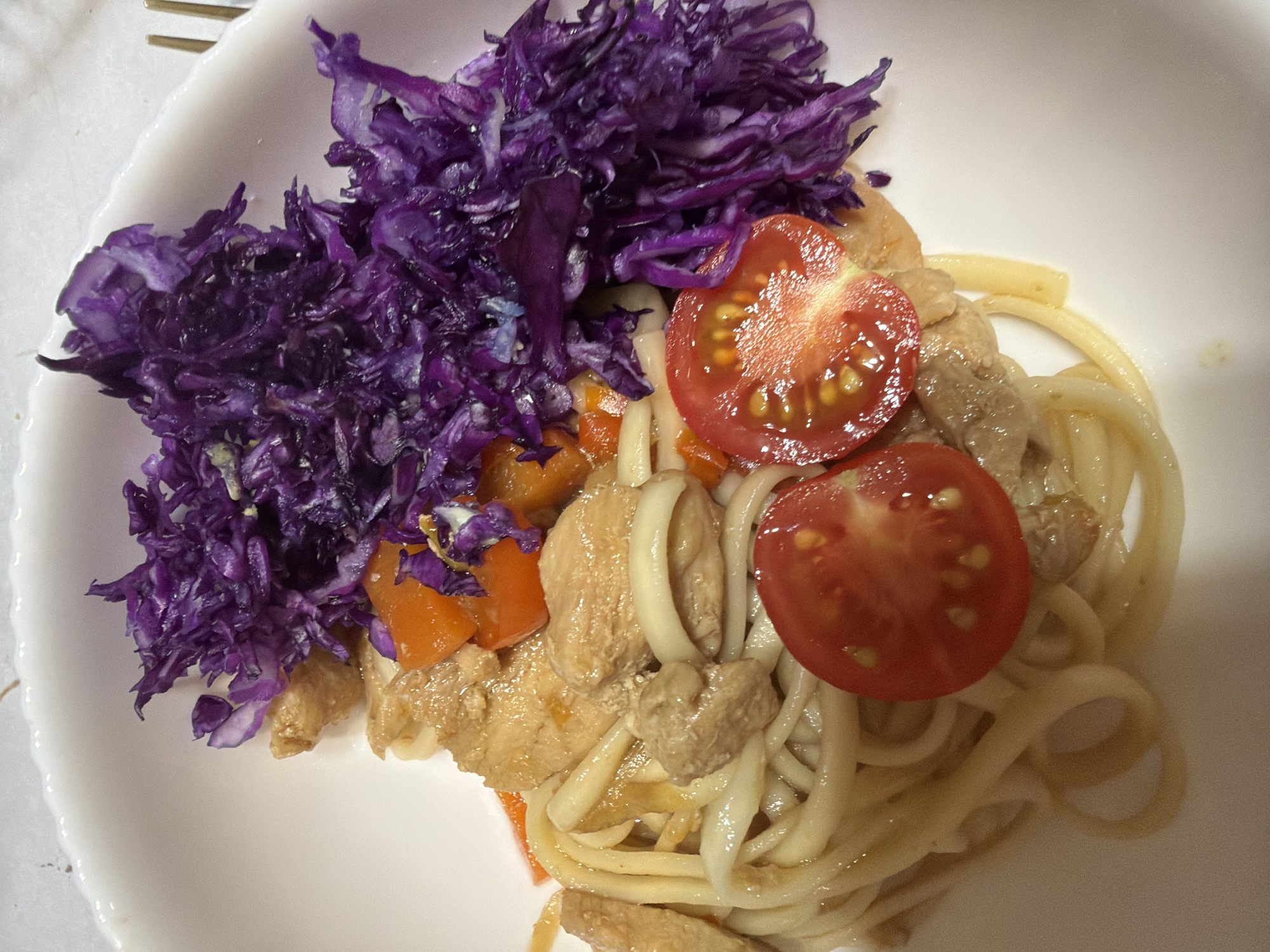 This close-up, high-angle photograph captures a prepared meal served on a white, scalloped plate. The primary subjects are various food items, including a large mound of shredded purple cabbage on the left, offering a vibrant contrast. Alongside the cabbage, the main dish consists of thick, light-colored noodles (possibly udon), intermingled with pieces of cooked chicken and diced orange vegetables, likely carrots. Two bright red cherry tomatoes, cut in half to expose their seeds and pulp, garnish the dish on the right side. A metal fork is partially visible in the upper left corner, suggesting the meal is ready to be eaten.
The scene depicts an indoor setting, likely a home or casual dining environment in Chaikovsky, Russia, where this meal is being consumed or has just been served. No people are visible in the frame, nor is any discernible text present. The time of day and external weather conditions cannot be determined from the indoor lighting. The overall impression is a nutritious and colorful home-cooked dish.