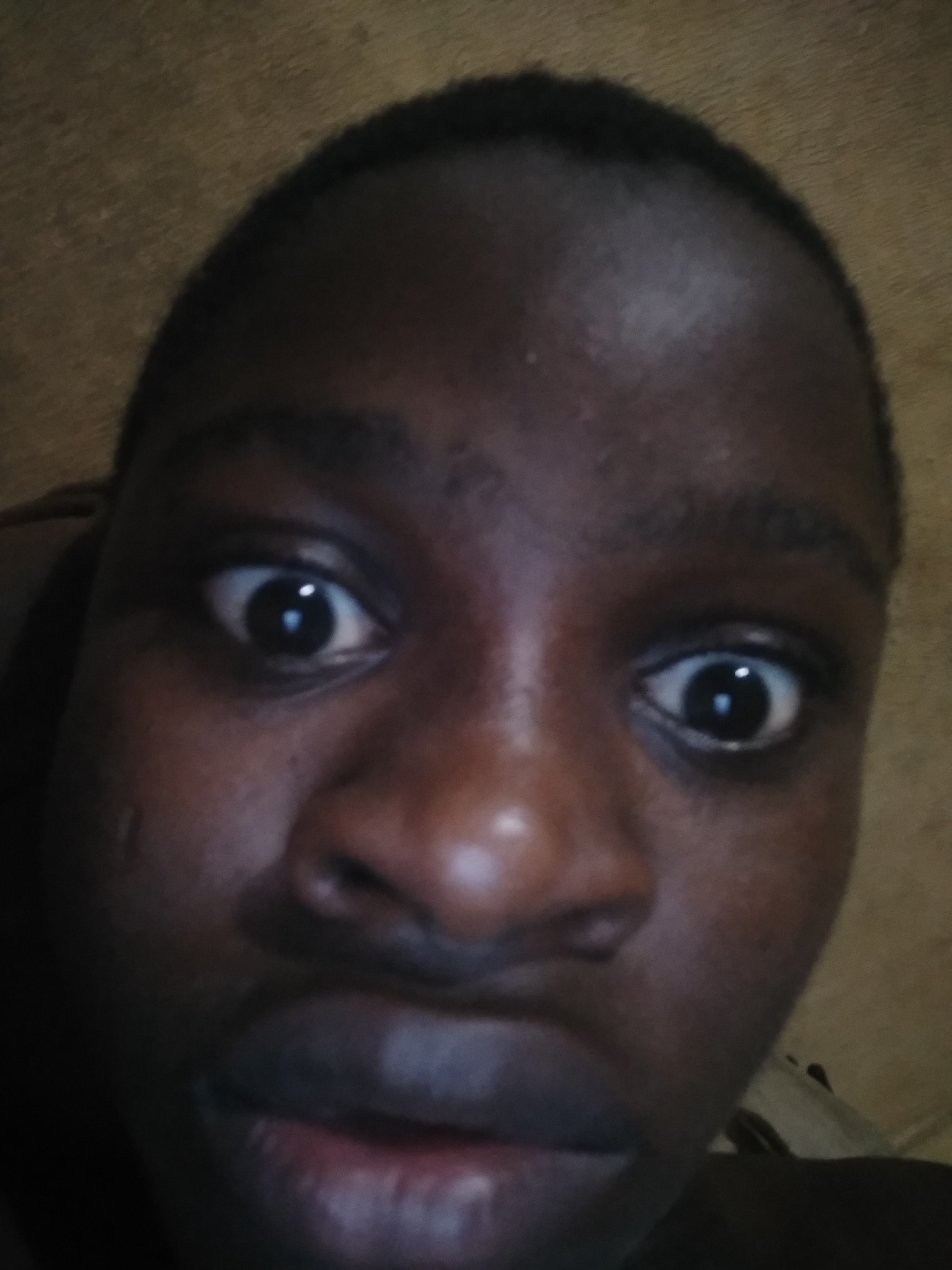A close-up, slightly low-angle selfie of a person with dark skin, wide eyes, and a serious expression. The background is indistinct and appears to be an indoor setting. The person is the sole subject in the frame. No specific location in any city is discernible.