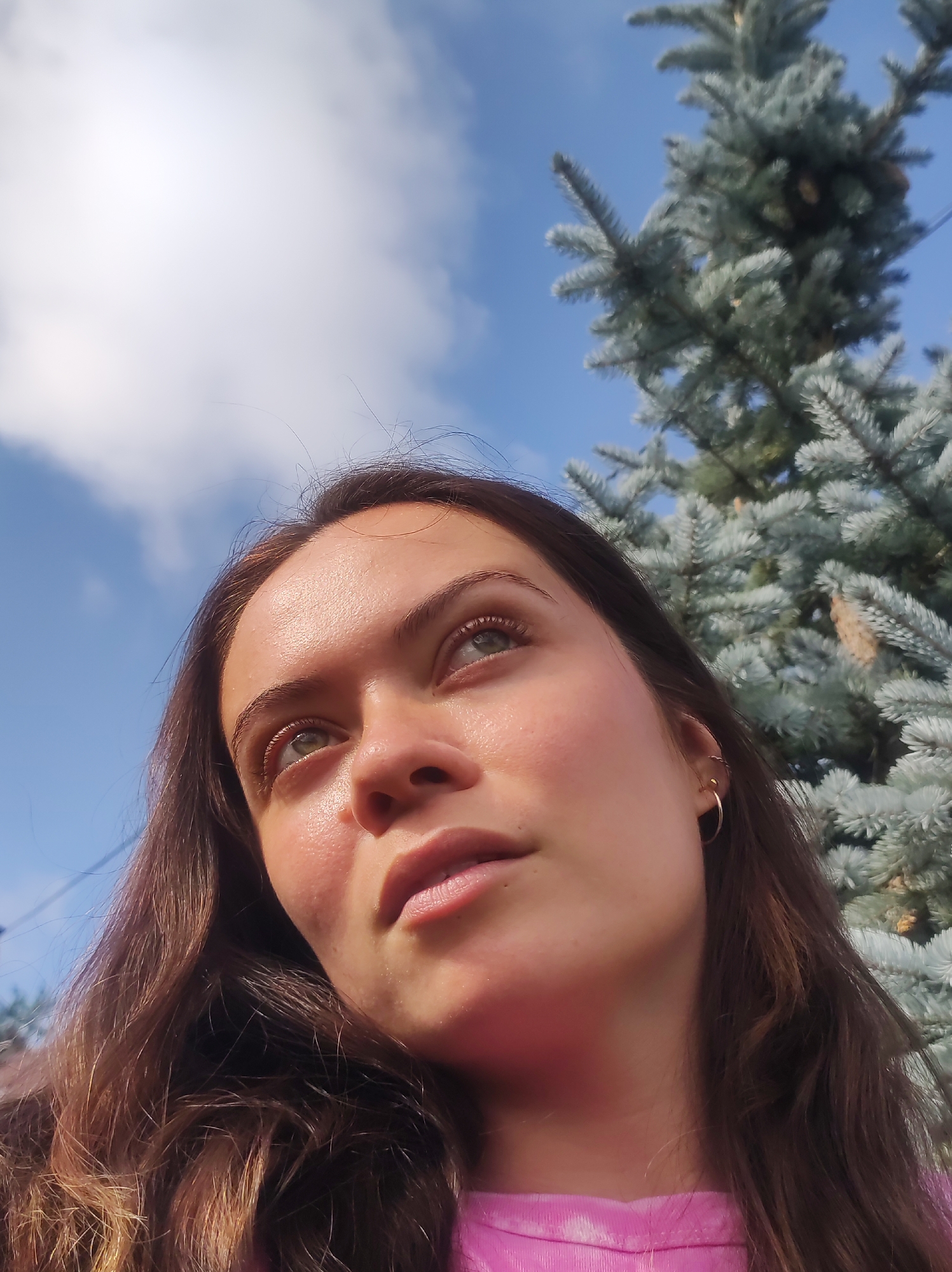 An outdoor, low-angle medium shot shows a young woman looking up. Her head is tilted back, and her eyes are directed towards the sky. She has long, brown, wavy hair that falls around her shoulders. Her skin is flushed, and she is wearing a pink shirt. To the right of the woman is a blue spruce tree. The sky is blue with scattered white clouds. The overall lighting suggests it is daytime, likely during sunny weather. The image was taken in the city of Fokino, Russia, likely in a park or residential area given the presence of a large tree.