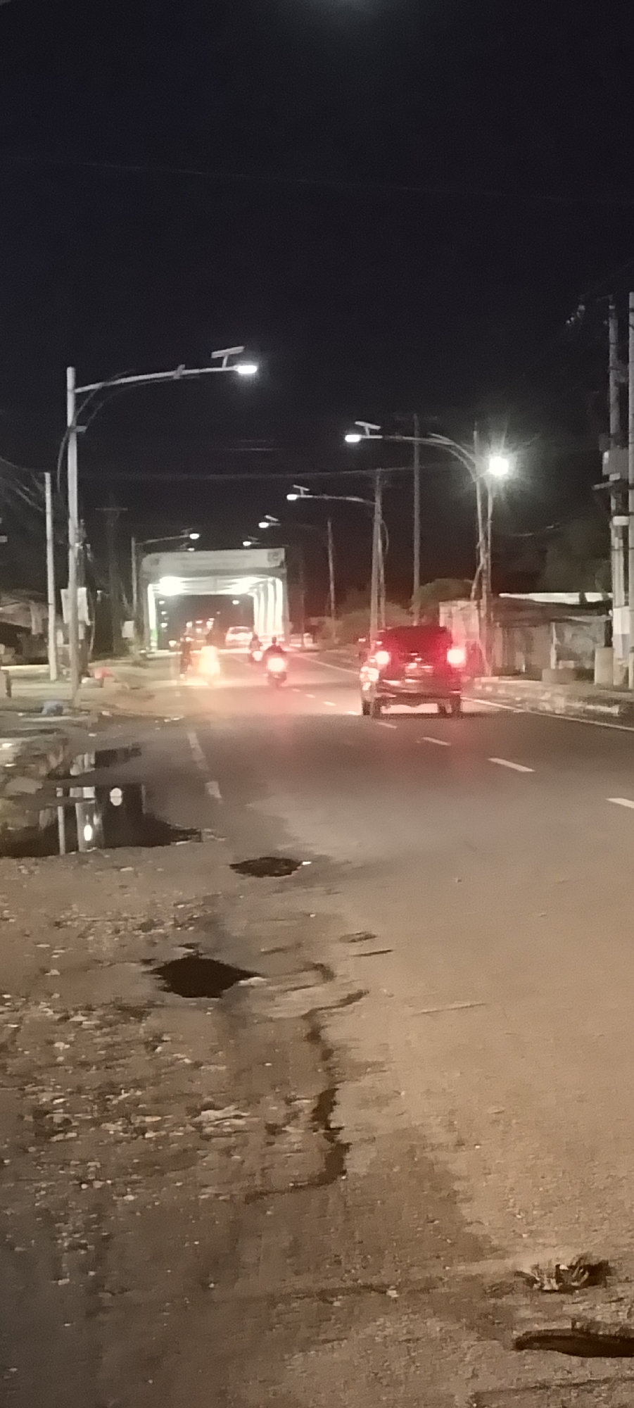 At night in Lhokseumawe, Indonesia, a road is lit by streetlights and vehicle headlights. The road surface is damaged and puddled. A dark-colored car drives away from the camera, its taillights illuminated. Two motorcycles, also with illuminated headlights, are further down the road near an illuminated overpass. Power lines run along the right side of the road. The night sky is dark.