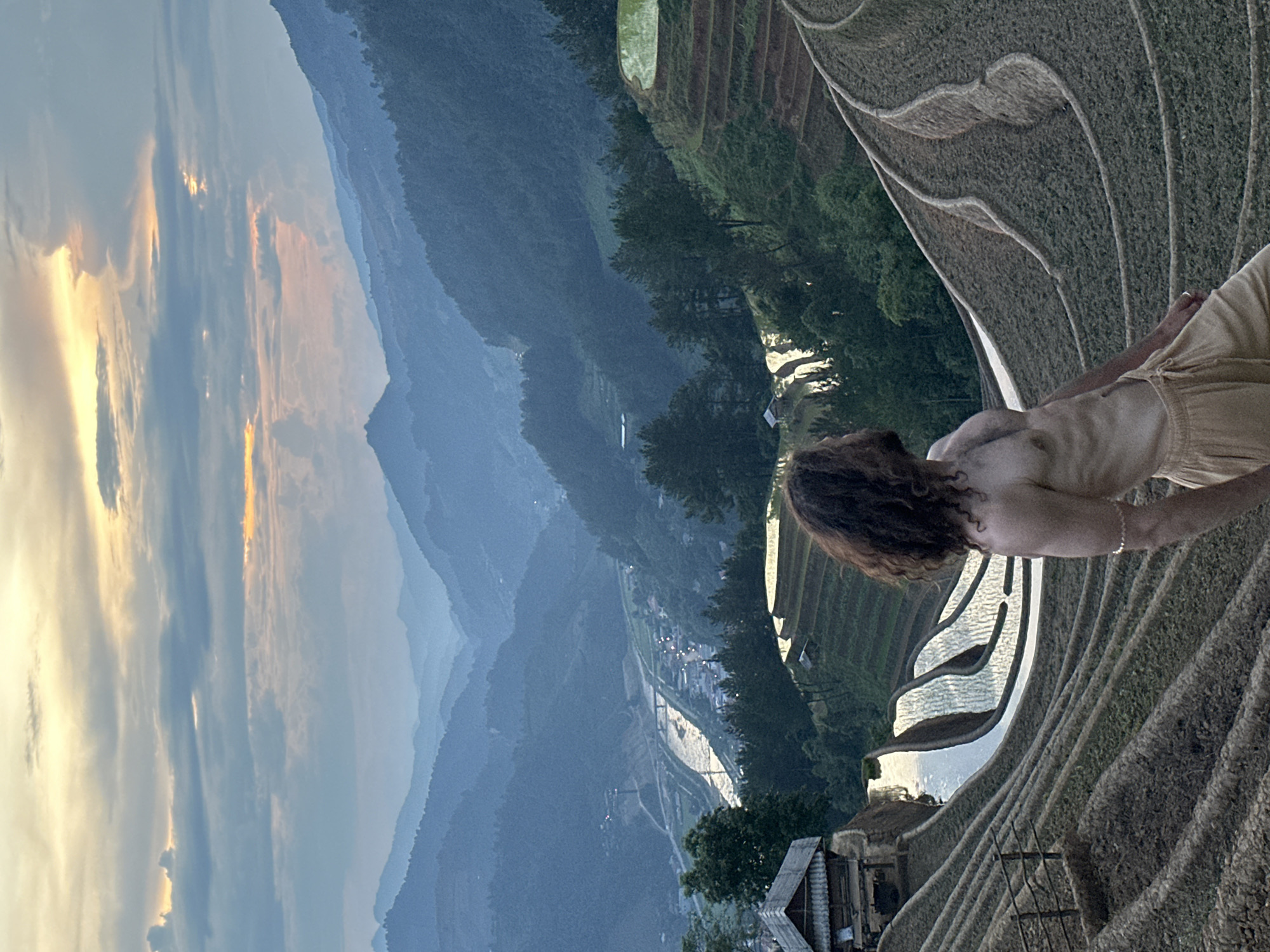 The image presents a panoramic landscape of terraced rice fields and mountains in Mù Cang Chải, Vietnam, captured at sunset or dusk. A person, viewed from behind, stands on the right side of the frame, facing away from the camera and observing the vast scenery. This individual has long, dark, wavy hair and wears light-colored, loose-fitting clothing, with a bracelet visible on their right wrist.
The foreground and mid-ground are defined by the intricate patterns of terraced rice fields. Many terraces are filled with water, beautifully reflecting the vibrant hues of the sunset sky—golden oranges, soft yellows, and muted blues—creating striking shimmering lines across the landscape. Further into the mid-ground, layers of rolling mountains emerge, some densely covered in dark green trees, while others in the distance appear softer and hazy. A valley winds through these mountains on the left side, where scattered buildings or a small village are visible, marked by faint lights that illuminate the approaching evening. A rustic building is partially visible in the lower right foreground, nestled within the terraced environment.
The activity depicted is the quiet contemplation or appreciation of the majestic natural beauty. The time of day is clearly sunset or dusk, indicated by the rich colors in the sky and the emerging lights in the valley. The atmosphere is one of serene grandeur. There is no visible text in the image.