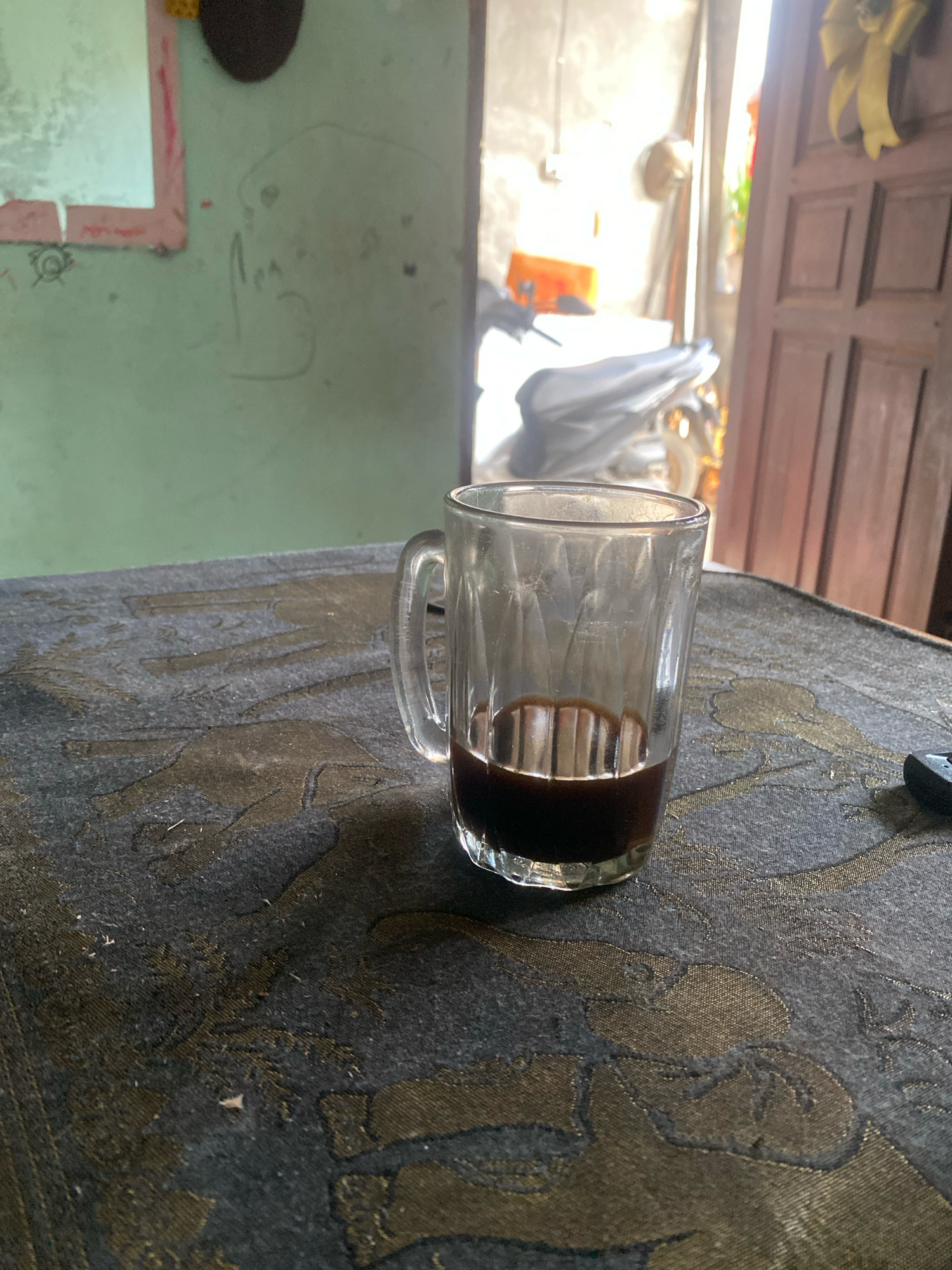 A glass of dark liquid sits on a patterned cloth-covered surface. The glass is clear and appears to be a shot glass or small tumbler. The liquid inside is dark brown, suggesting coffee or a similar beverage. The cloth has a dark background with a metallic gold pattern that appears to depict abstract shapes or possibly animals. In the background, a motorcycle is partially visible, along with a doorway and a portion of a wooden door. The wall in the background is painted a light green color and has some faint markings on it. Sunlight is entering from the right side of the image, creating highlights and shadows. The scene appears to be an indoor setting, possibly a home or a small establishment. This location is in Batam, Indonesia.