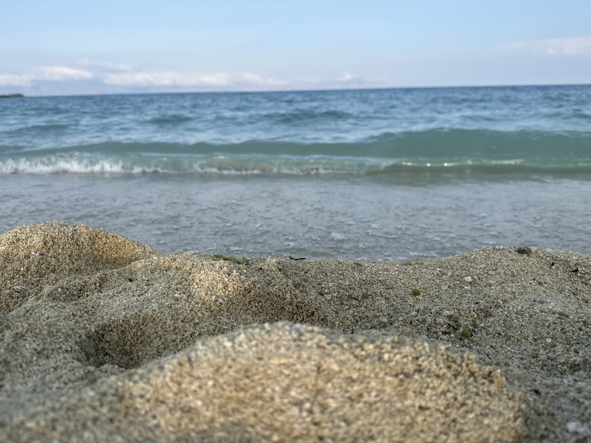 A low-angle, close-up shot shows a sandy beach in the foreground with gentle waves washing ashore from a calm, blue sea in the background. The sky above is clear and pale blue with faint clouds on the horizon. The sand is light brown and textured, with small pebbles and scattered bits of debris. A small amount of green sea grass is visible in the sand. The water is clear and a vibrant turquoise near the shore, deepening to a dark blue further out. The image is sharp and detailed, focusing on the textures of the sand and the movement of the water. The overall impression is one of a peaceful and serene beach scene. There are no people or man-made objects visible. The location is not identified as Norashen, Armenia, but rather is a generic beach.