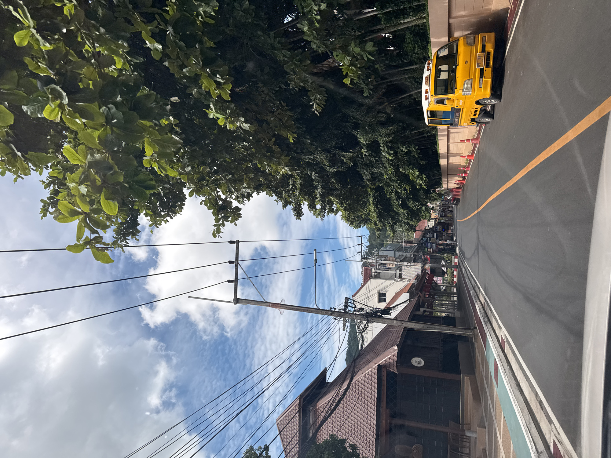The image depicts a street scene in a city, likely in a tropical or subtropical region, given the lush green foliage and the bright, sunny sky with scattered clouds. The street is paved and has a yellow line marking the center. On the left side of the street, there is a building with a traditional-looking, multi-tiered, brown tiled roof and dark wooden walls. Adjacent to it are other buildings with awnings and signs, suggesting a commercial area. A utility pole with numerous wires stretches across the sky. On the right side of the street, a small, bright yellow taxi truck is parked parallel to a light-brown, textured wall. Orange traffic cones are placed along the edge of the road near the taxi. Tall, leafy trees line the right side of the street behind the wall, with palm trees visible interspersed among them. The overall atmosphere is that of a daytime scene in an urban environment that incorporates natural elements.